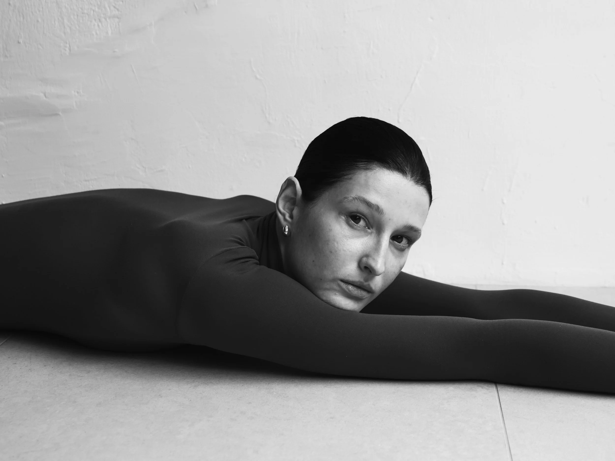 A woman with dark hair slicked back, wearing earrings, is lying on her side on the floor with her head resting on her arm, in a black-and-white photo with a plain wall background.