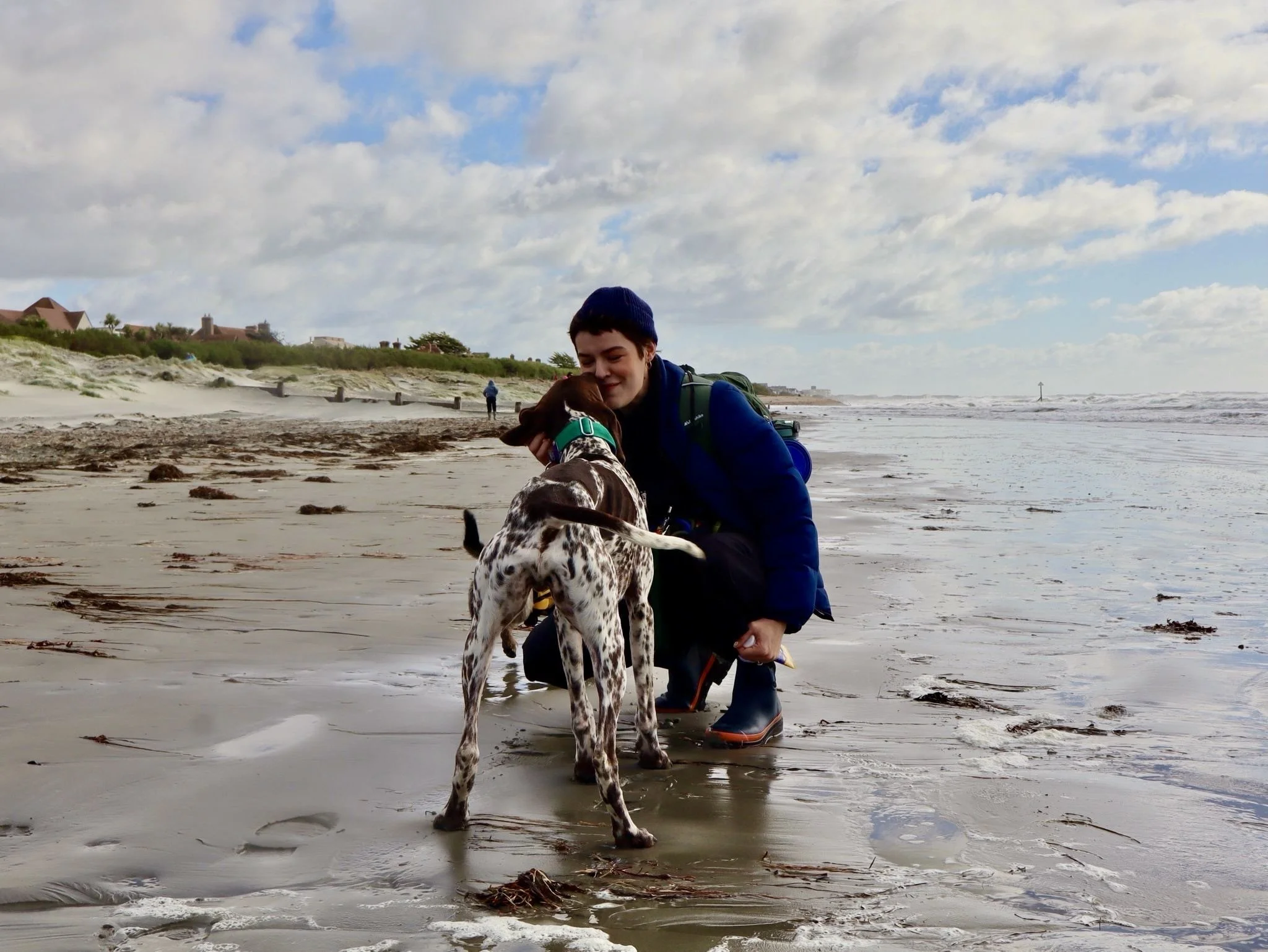 A person with short hair, wearing a navy blue jacket, kneeling on a sandy beach, hugging a Dalmatian dog with a green collar near the shoreline during daytime.