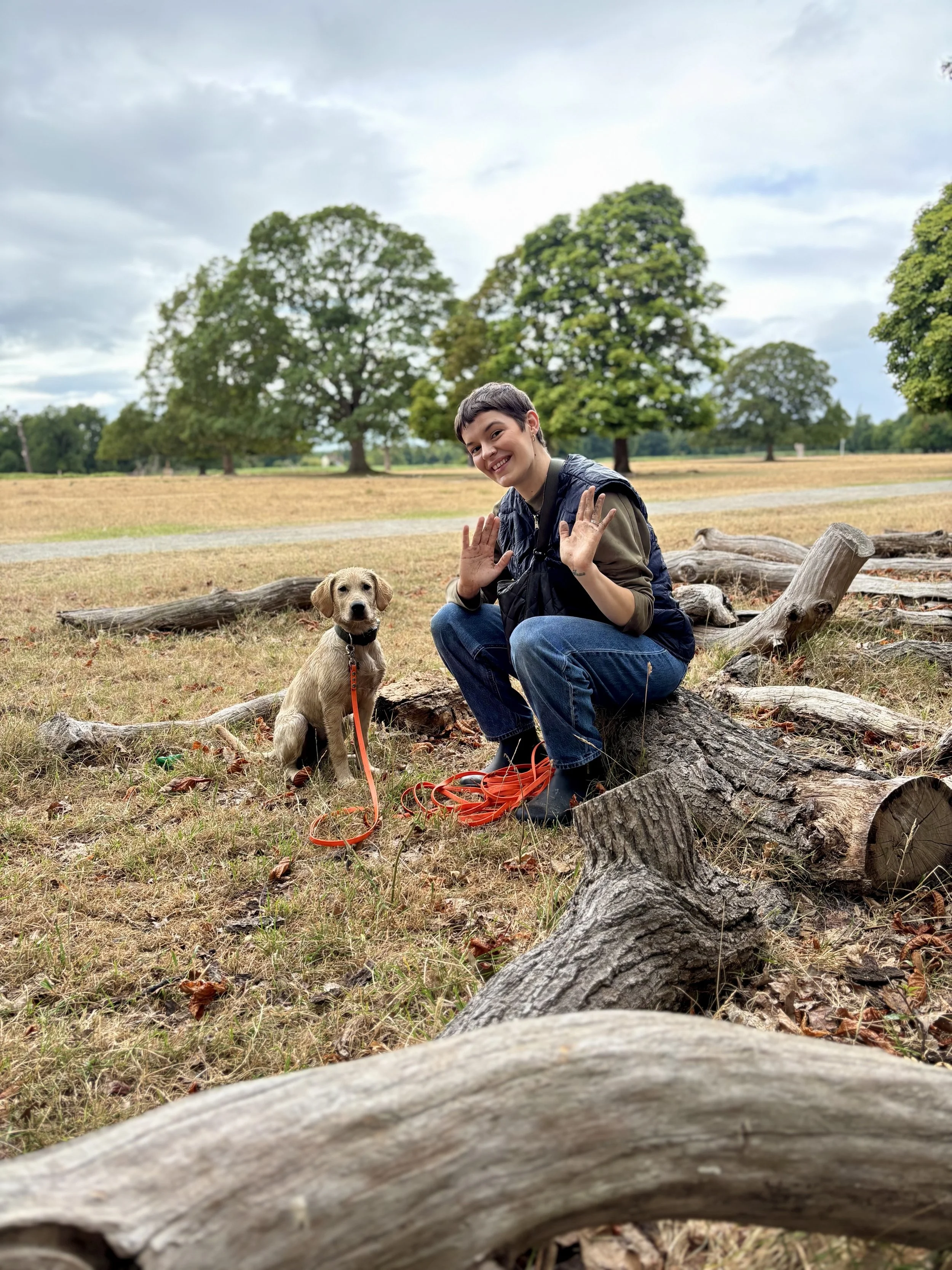 A young person with short dark hair smiling and raising their hands, sitting on a log with a tan puppy on a leash in a park with trees and a cloudy sky in the background.