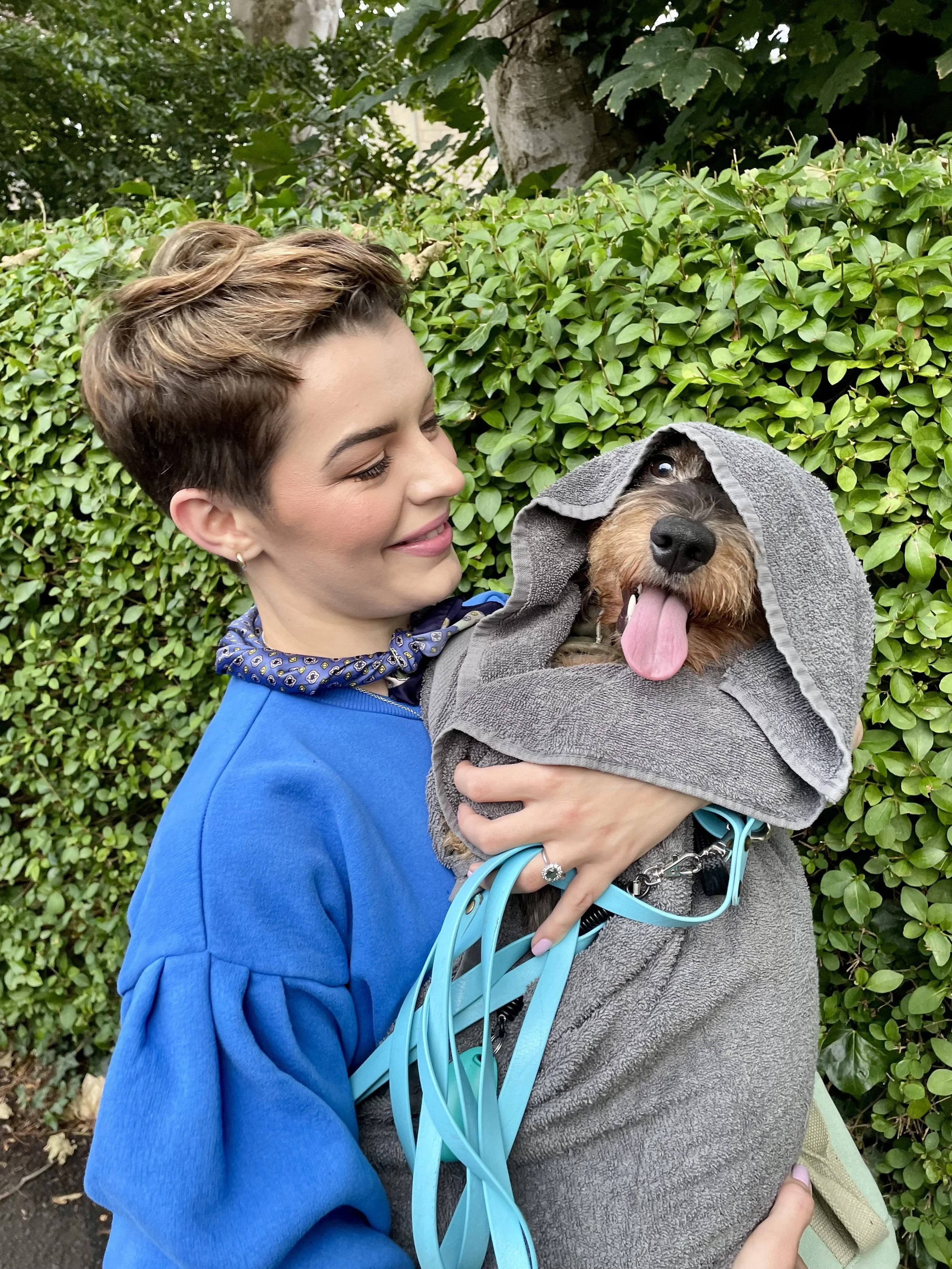 A woman holding a dog wrapped in a grey towel, the dog has its tongue out and is partially covered with a hood, with a green bush and some trees in the background.