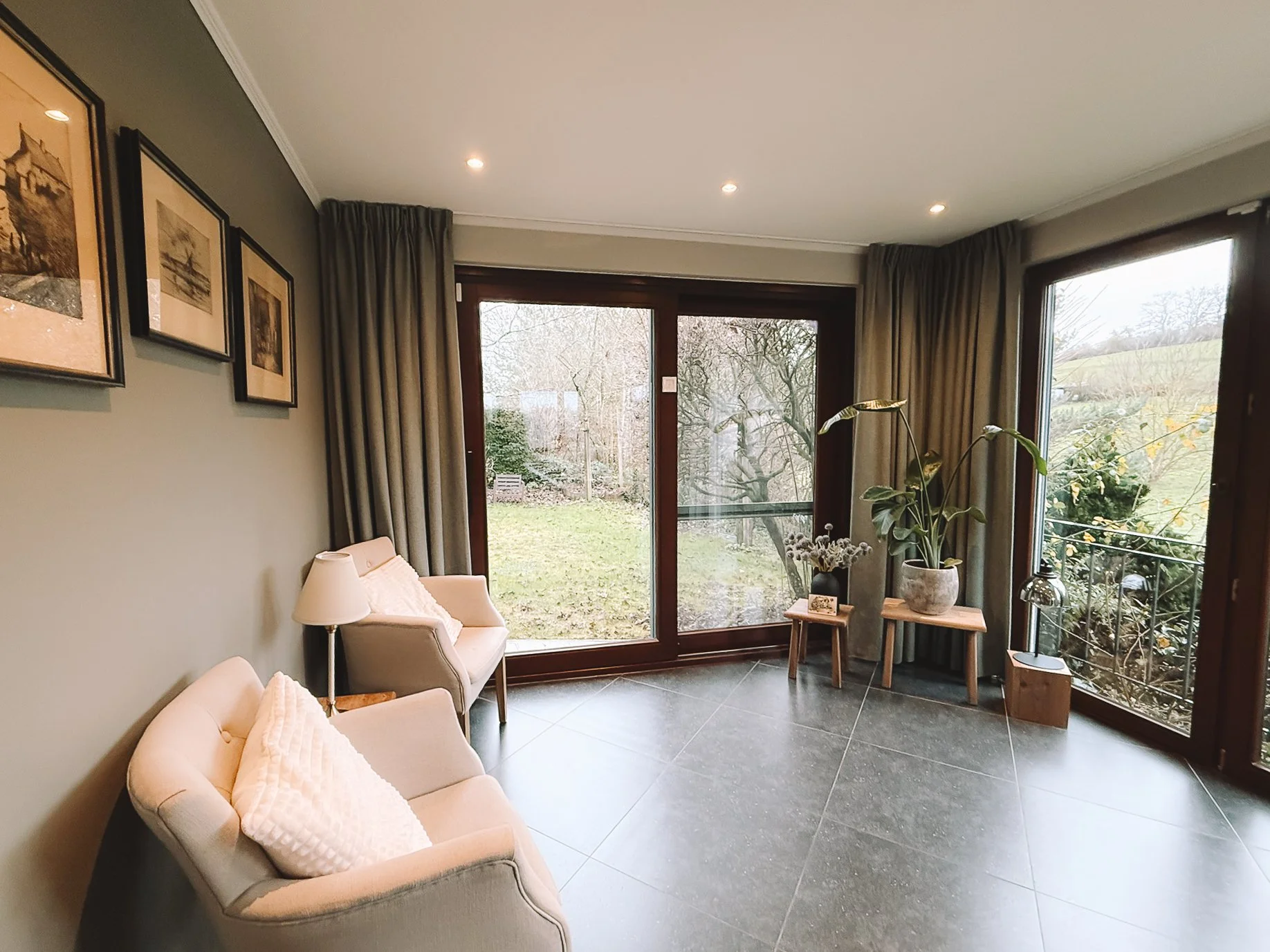 Woonkamer met grote ramen, planten en zithoek met twee fauteuils en een vloerlamp.