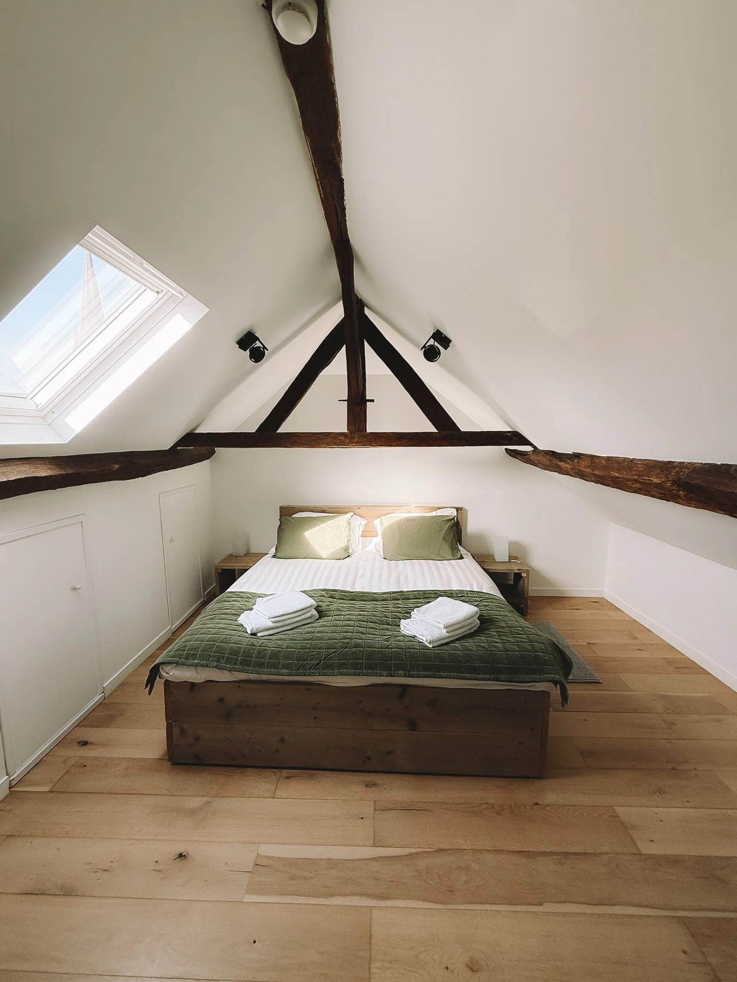 Een slaapkamer met een bed onder een gewelfde, gebinten plafond met een dakkapel aan de linkerkant en twee nachtkastjes.