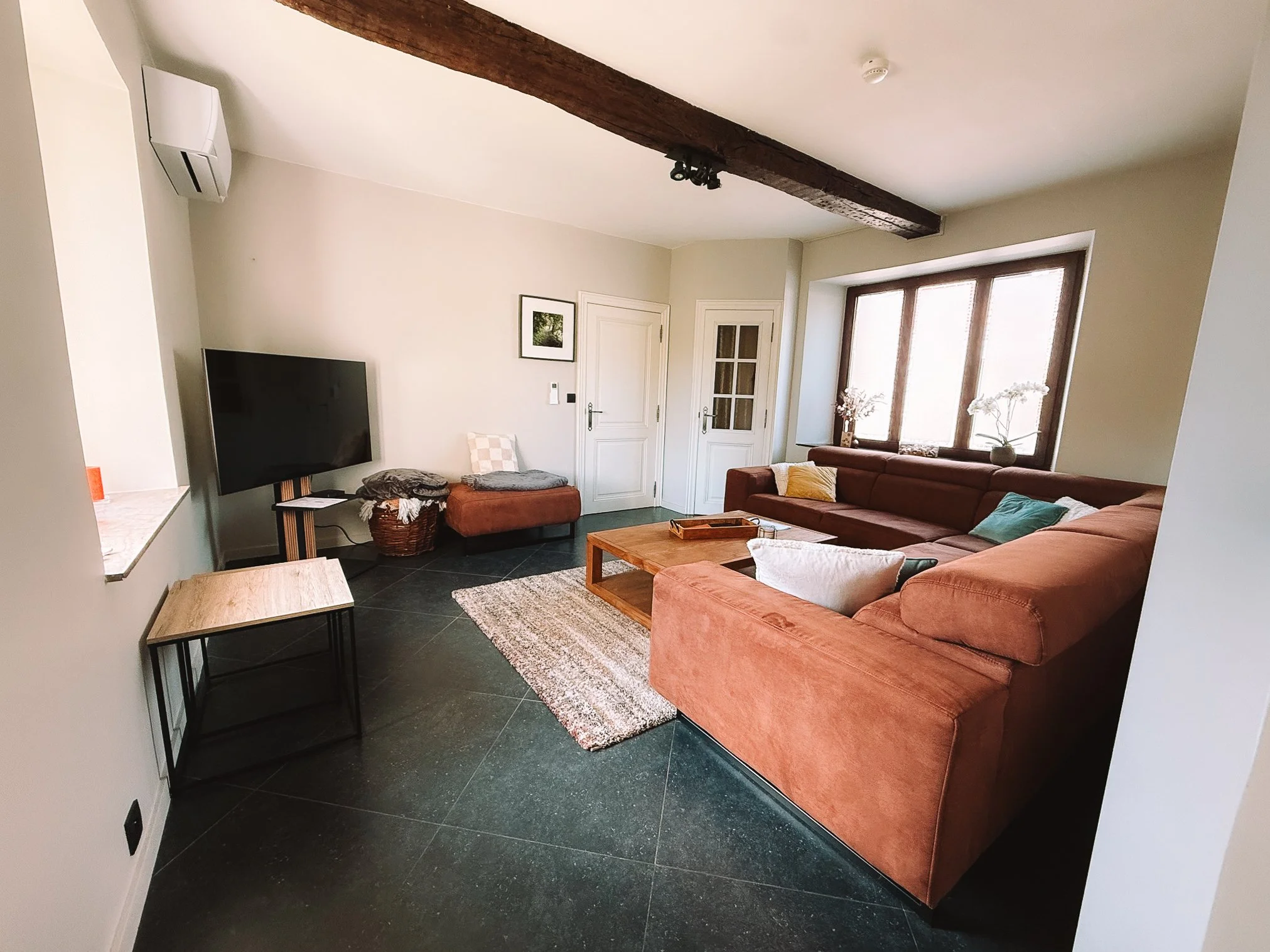 Woonkamer van het vakantiehuis Au bord du Floyon met grote hoekbank, groot raam, tv aan de muur, houten salontafel en gestreept vloerkleed.