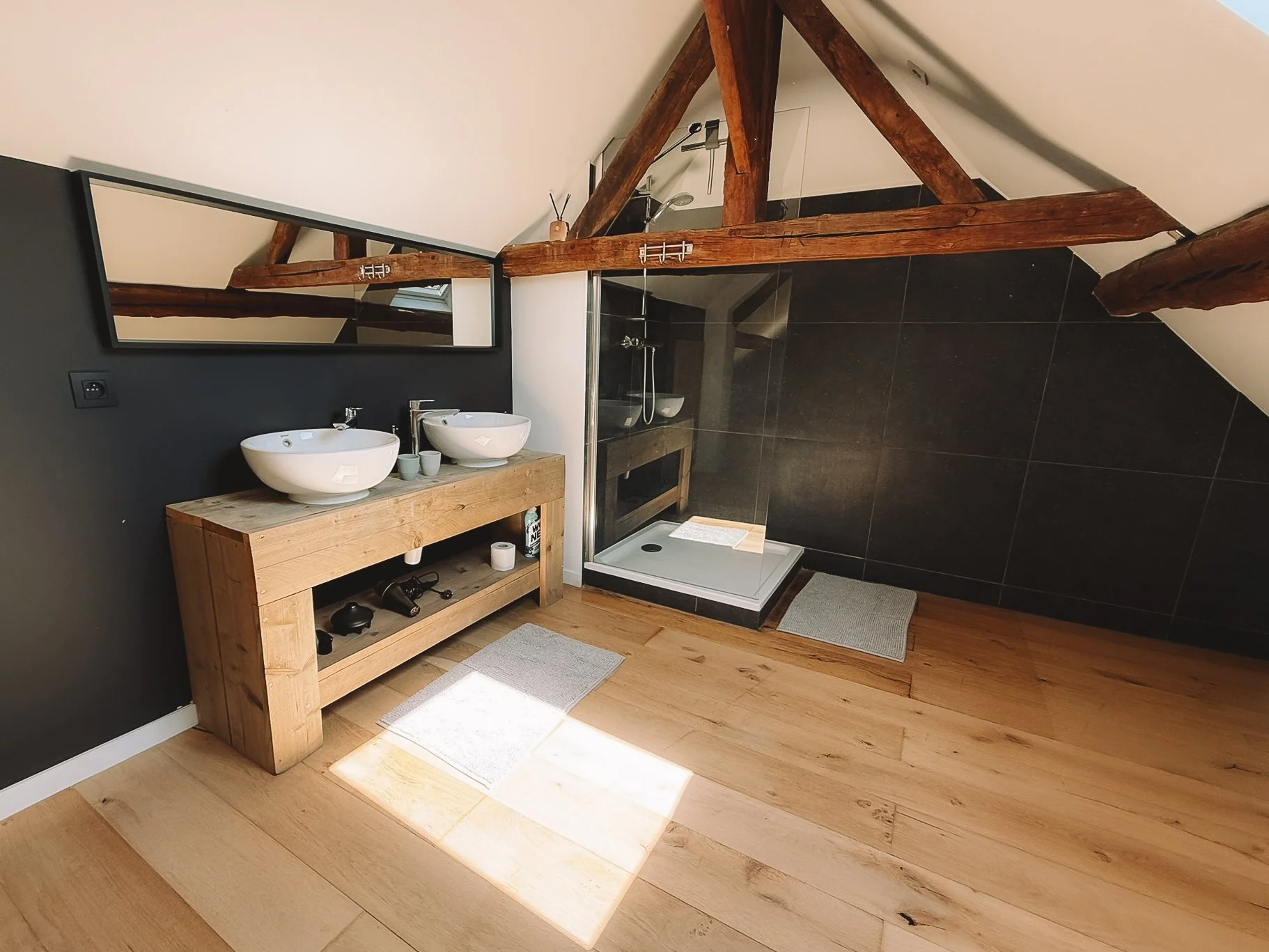 Benedenverdieping van een stijlvolle badkamer met twee waskommen op een houten meubel, een spiegel erboven, een inloopdouche, houten vloer en plafondbalken.