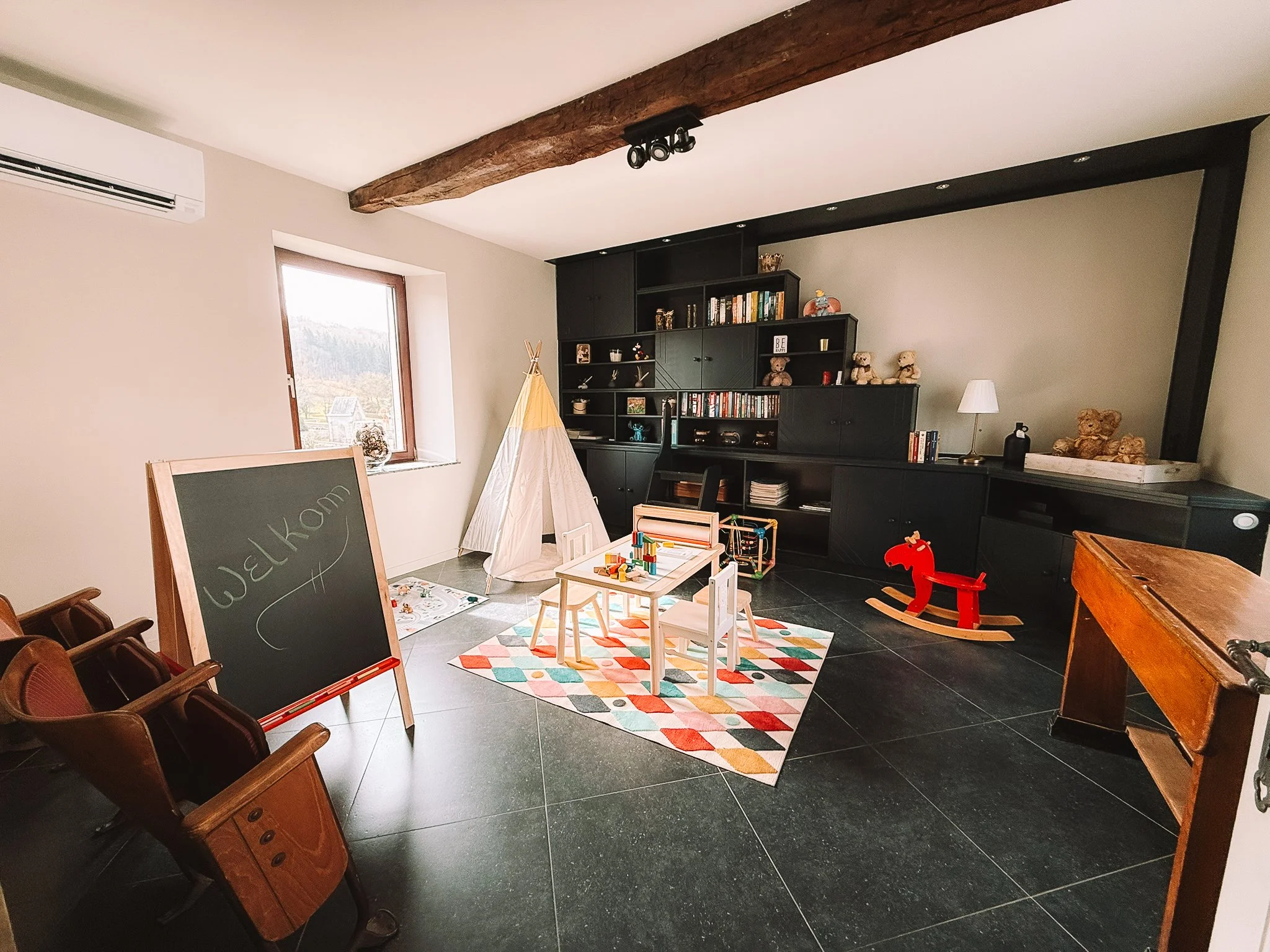Kinderkamer met speelkeukentje, tafeltje en stoelen, tipi tent en speelgoed, zwarte boekenkast met boeken en teddyberen, rode schommel in de vorm van een hond en een schoolbord met welkom opgeschreven.