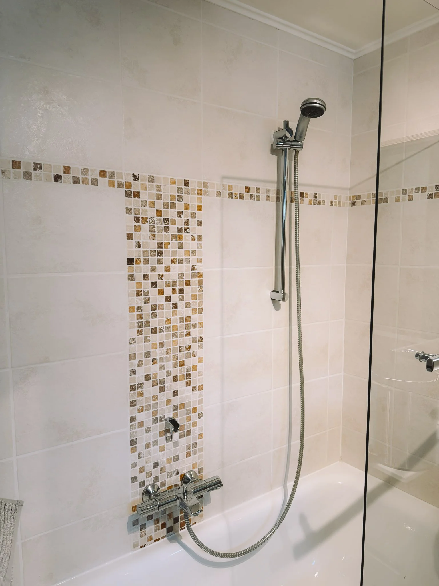 Badkamerdouche met een glazen scheidingswand, een handdouche, een muur met beige tegels en een decoratieve strook mosaïektegels.