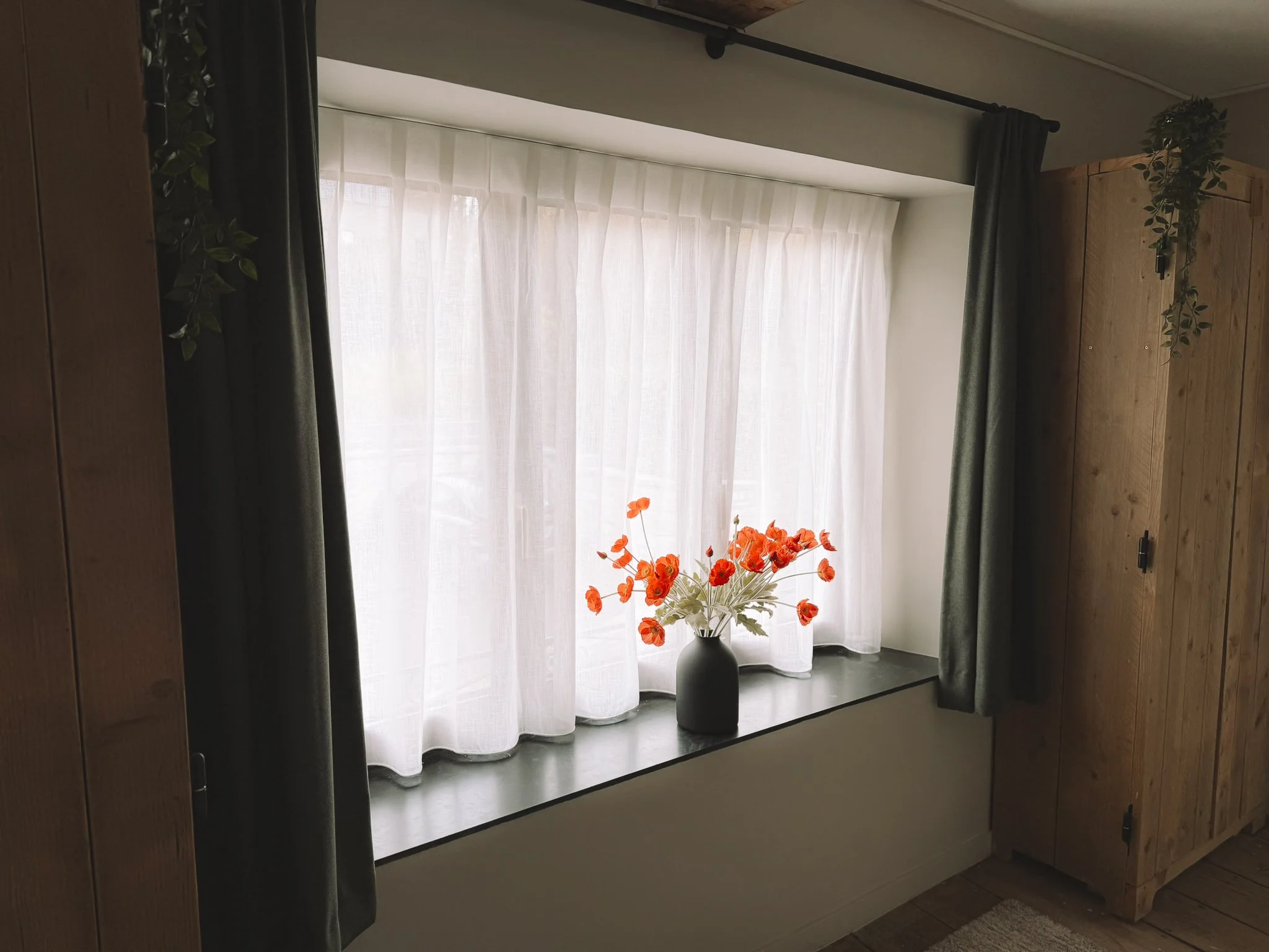 Een raam met witte gordijnen en een zwarte vaas met oranje bloemen.