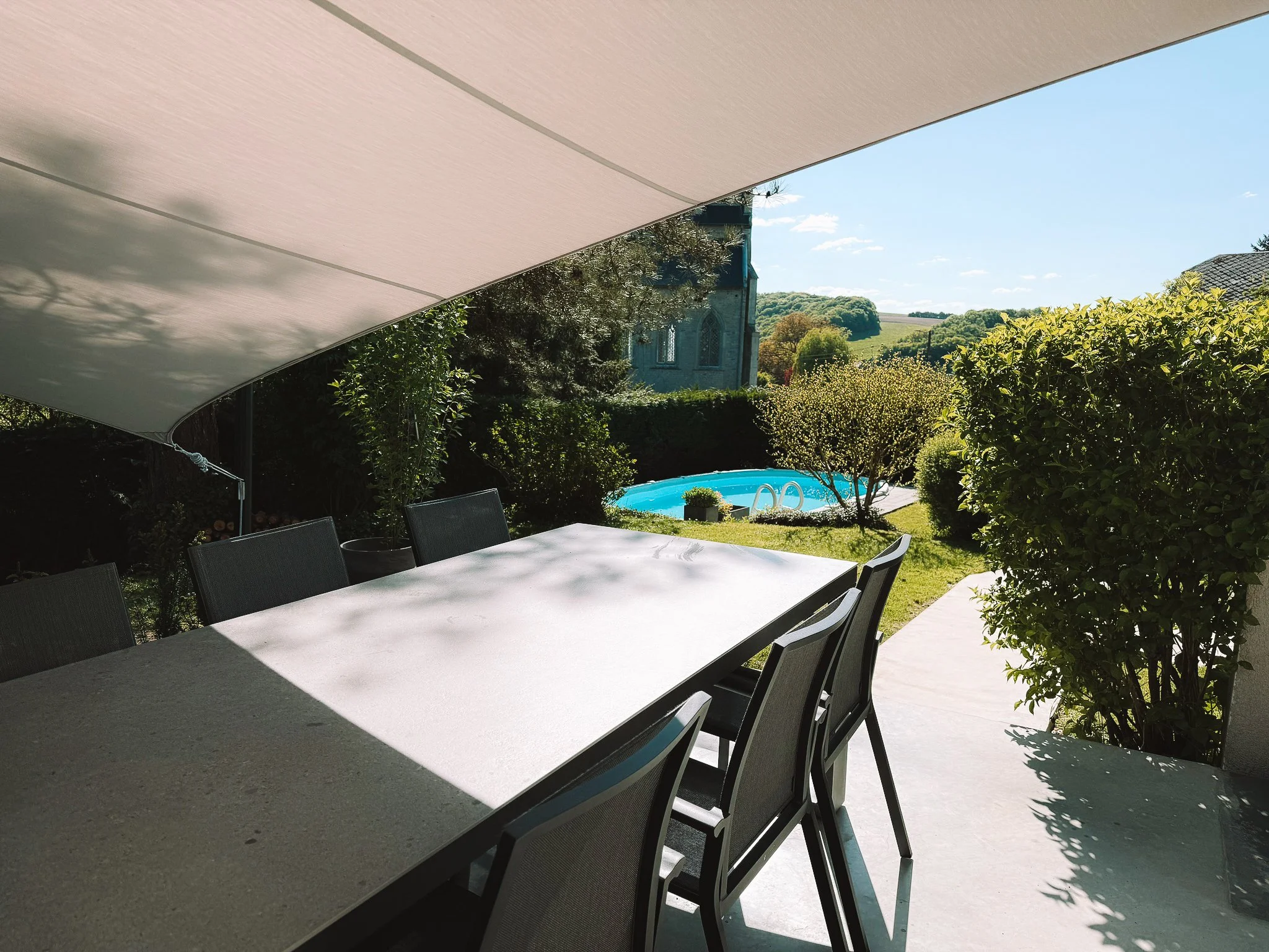 Buitenruimte met zwarte eettafel en een parasol, omgeven door groene struiken en een verwarmd zwembad.