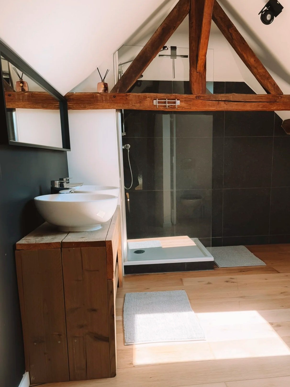 Een moderne badkamer met een houten kast, een ronde wastafel, een douche met zwarte tegels en een houten plafond met zichtbare balken.