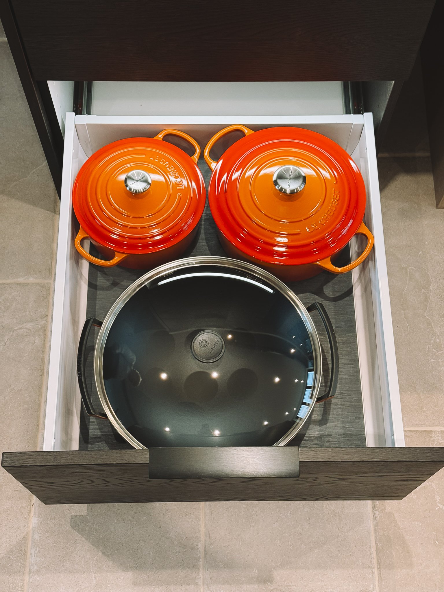 Twee oranje kleurrijke gietijzeren potten met deksels en een grote metalen pan, allemaal in een lade onder een kastenwand.