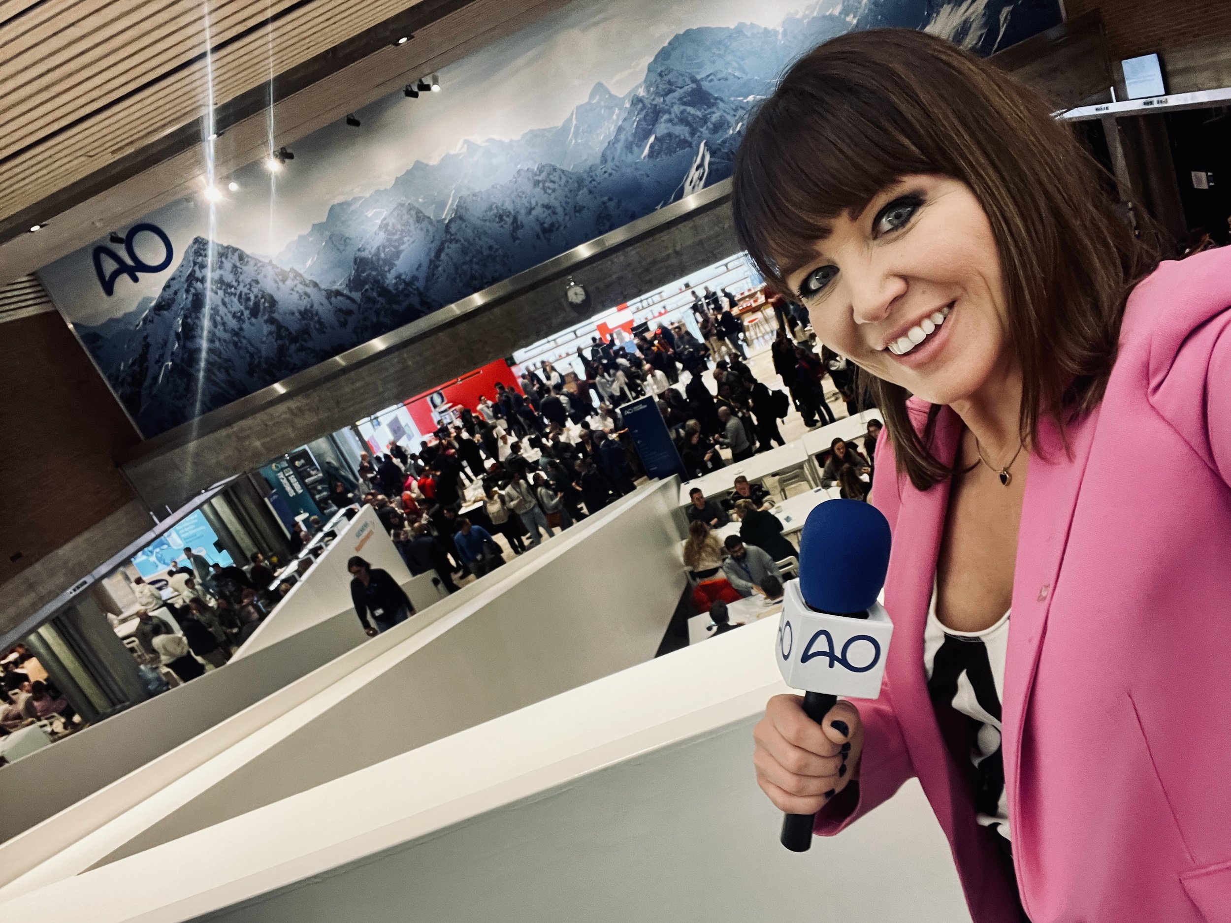 A woman with brown hair, wearing a pink blazer and holding a microphone, takes a selfie inside an airport with a large crowd of travelers in the background. The airport features a mural of snowy mountain peaks and the airport code 'AO'.