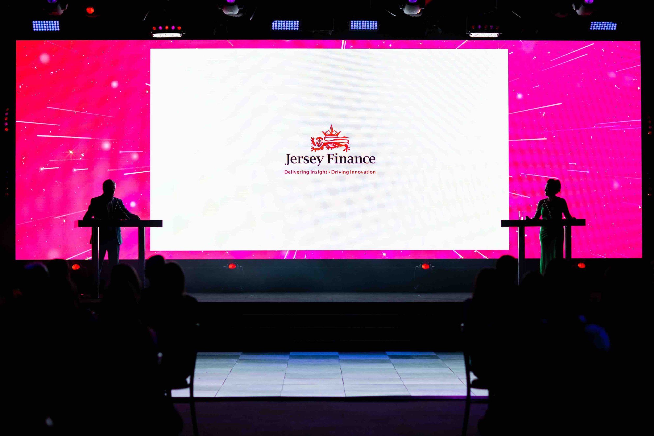 A stage with a large screen displaying the logo and name of Jersey Finance, with the tagline "Delivering Insight. Driving Innovation." Two silhouetted presenters stand at podiums on either side of the stage, with an audience watching in the foregroun