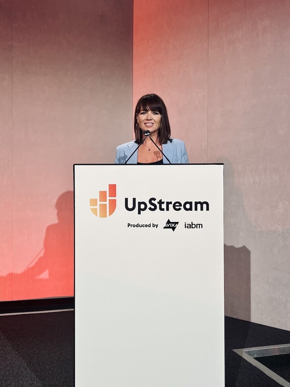 A woman standing behind a podium giving a speech at the UpStream event. The podium features the UpStream logo and it is produced by AVIXA and IABM.