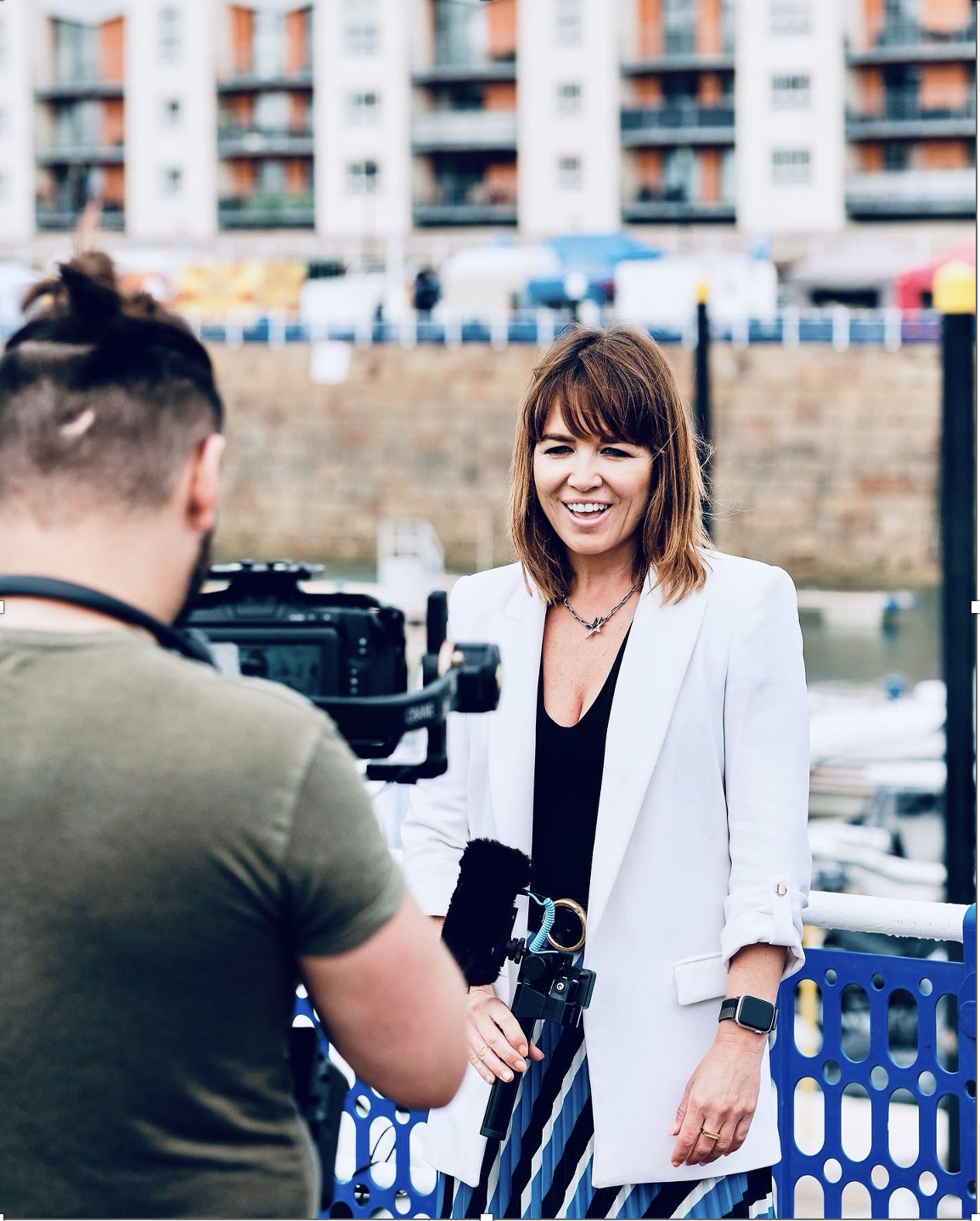A woman with shoulder-length brown hair and a white blazer is being filmed by a man operating a camera near a waterfront with buildings and boats in the background.