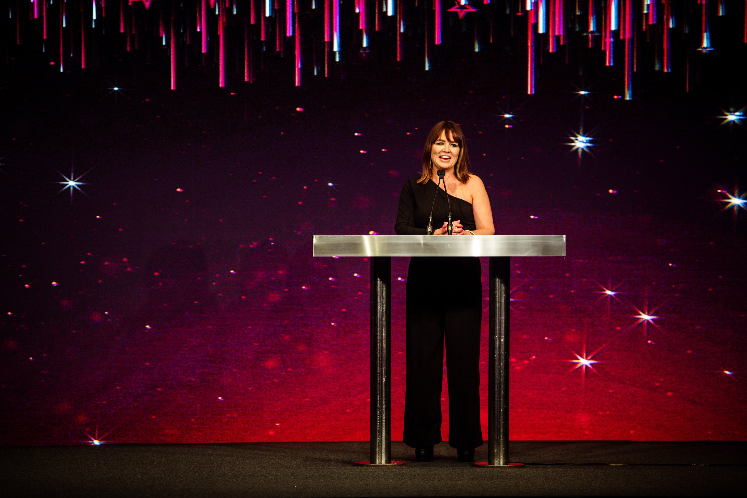 A woman in a black dress speaking at a podium on a stage with a colorful, starry background.