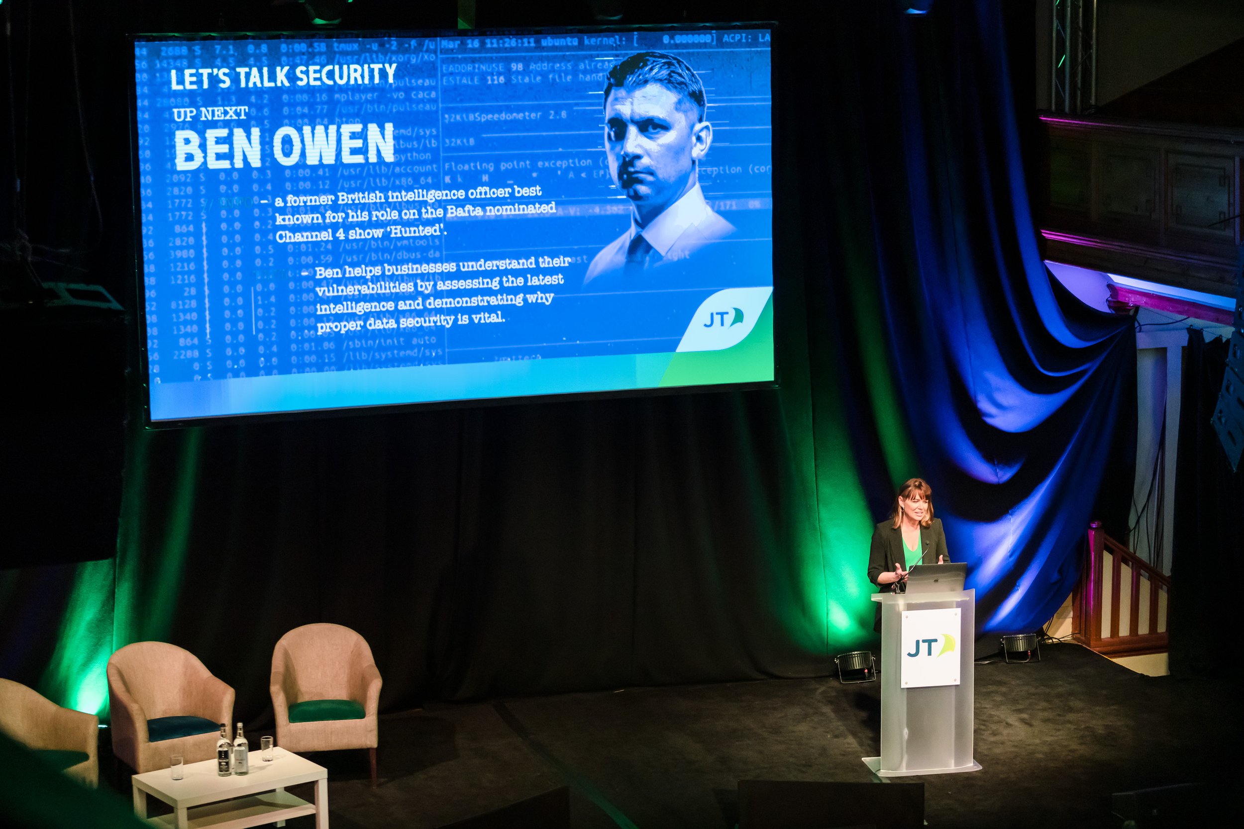 A woman standing at a podium giving a presentation on stage with a large screen behind her displaying information about Ben Owen, a British intelligence officer, with chairs and a small table in the foreground.