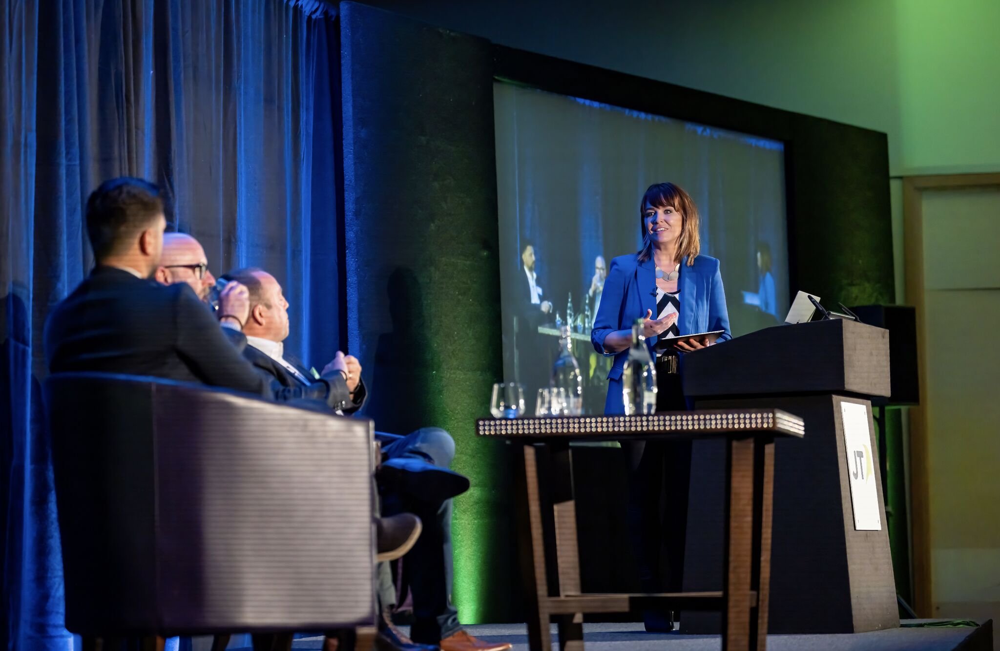 A woman in a blue blazer is speaking at a panel discussion with three men seated on the stage, while a large screen behind them displays her image, and water bottles are on the table in front of her.
