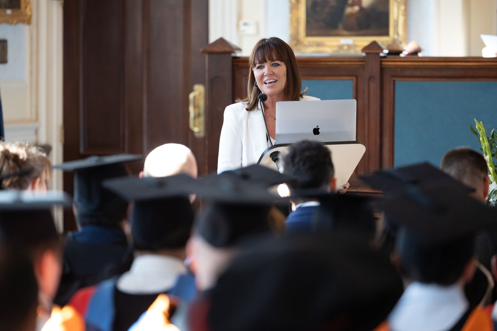 Woman giving a lecture at a graduation ceremony, standing behind a podium with a laptop, speaking to students wearing caps and gowns.