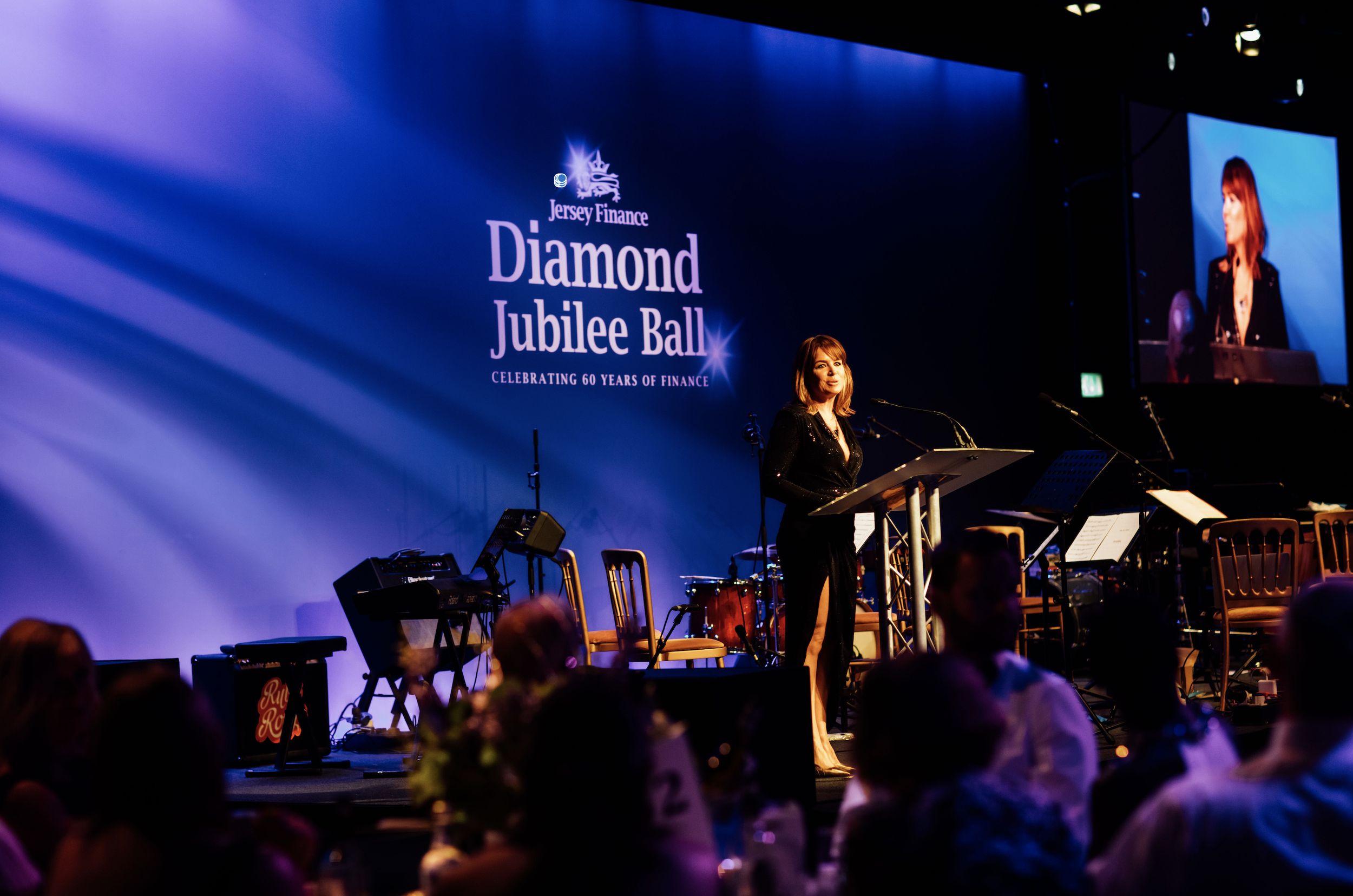 A woman in a black dress stands at a podium speaking at the Jersey Finance Diamond Jubilee Ball, celebrating 60 years of finance, with a large screen behind her displaying the event logo and a live feed.