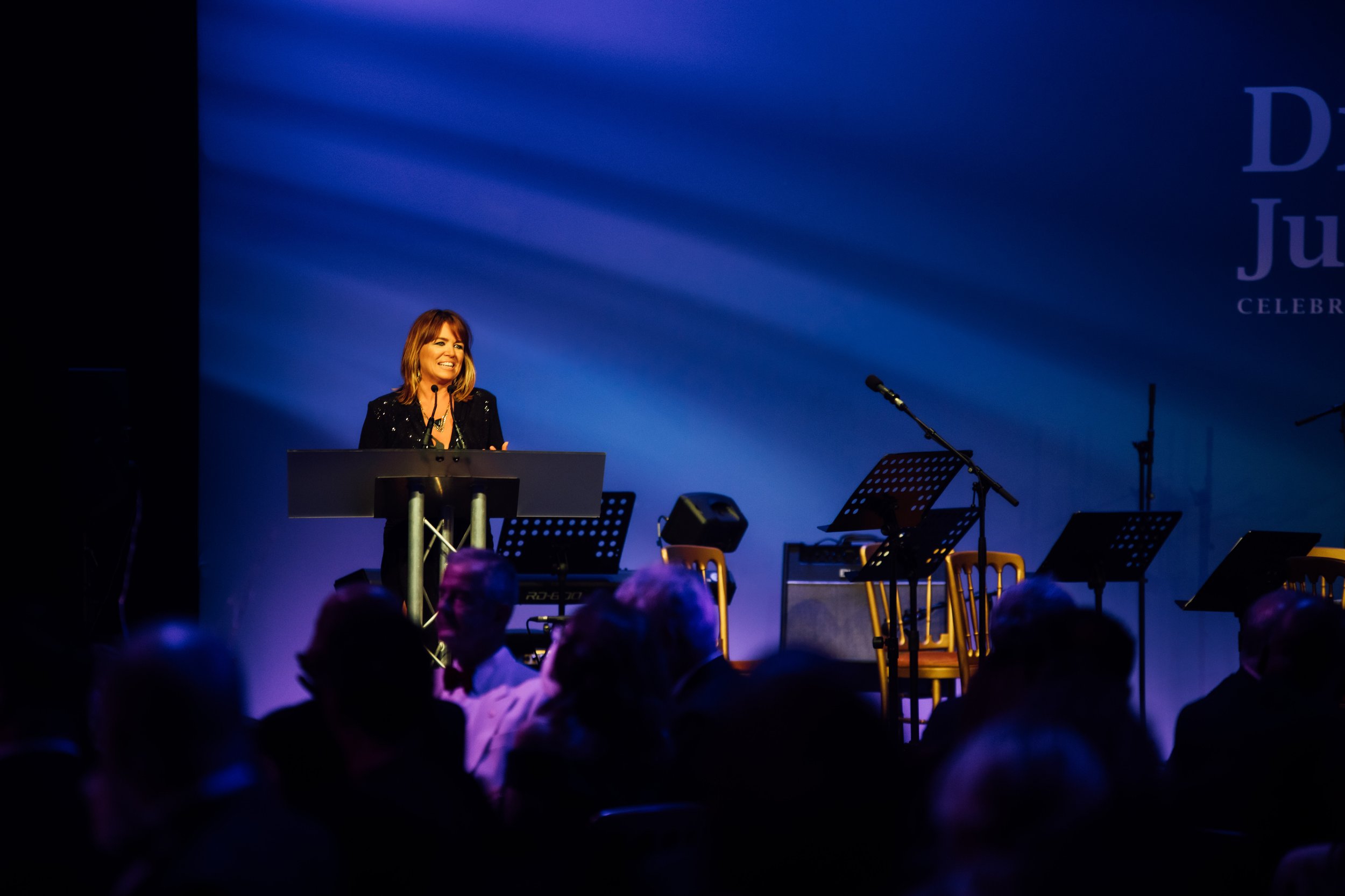 A woman with shoulder-length hair speaking at a podium during an event on a stage with musical instruments, illuminated by stage lights, with an audience in the foreground.