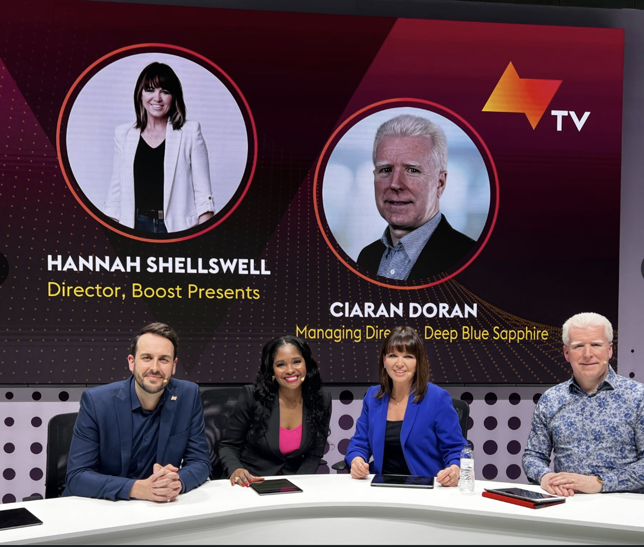 Television studio with four people seated at a white table and a large screen behind them showing portraits of Hannah Shellswell and Ciaran Doran. Hannah Shellswell is labeled as Director, Boost Presents; Ciaran Doran as Managing Director of Deep Blu