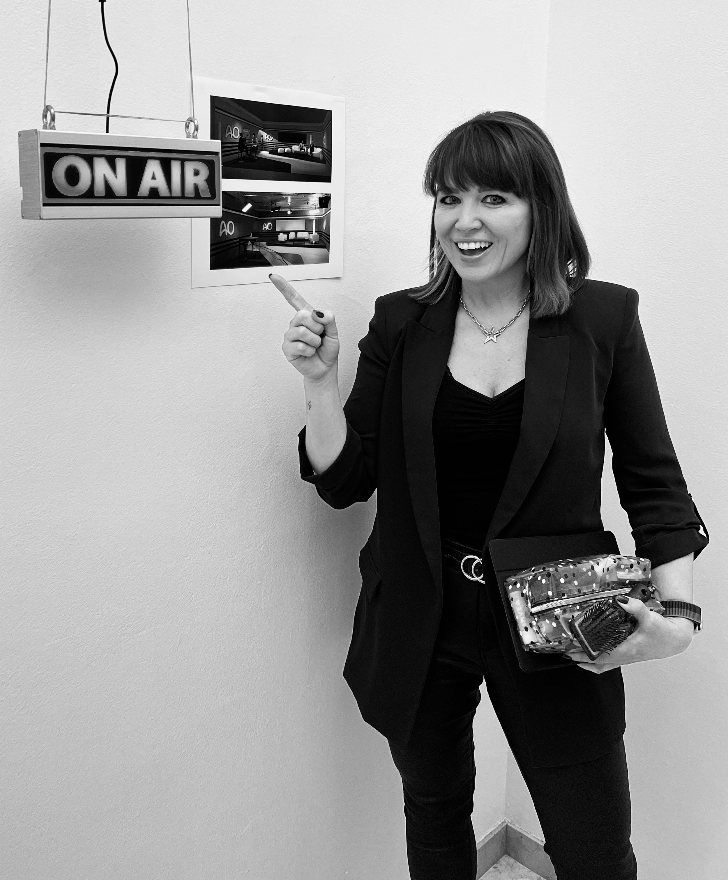 Woman smiling and pointing at a wall with two monitors and a sign that says 'ON AIR' in a studio or office setting.