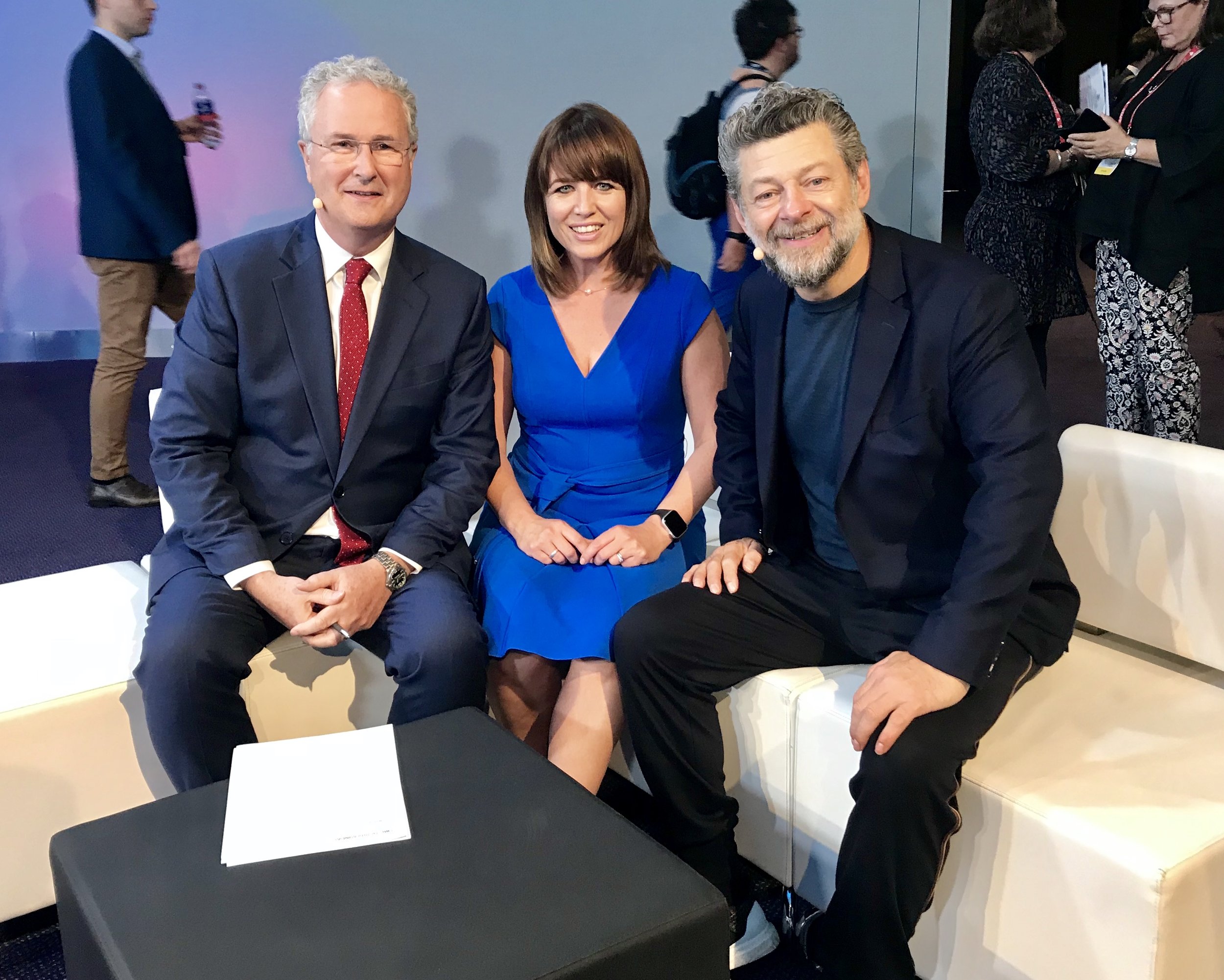 Three people sitting on a white sofa, smiling at the camera, in a conference or event setting. The person on the left is a man in a suit and red tie, the person in the middle is a woman in a blue dress, and the person on the right is a man in a black