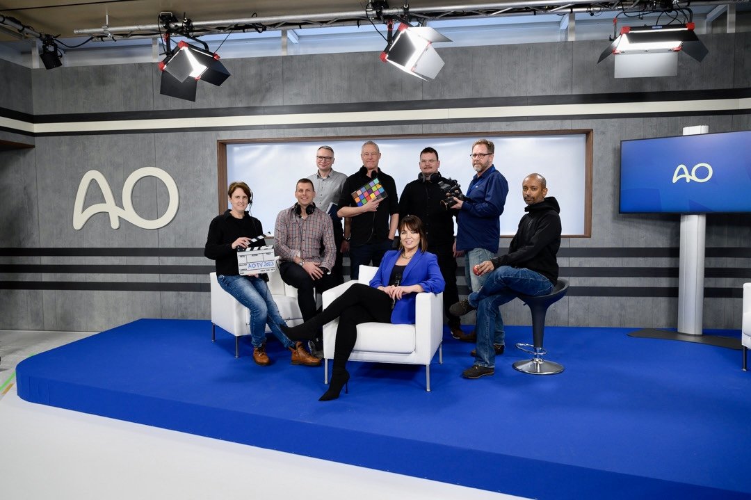 Group of eight people on a blue stage in a TV studio, with a woman in a blue blazer sitting on a white chair in front, and the others standing or sitting around her. Studio lights hang overhead, and AO logo is on the wall and a screen to the right.