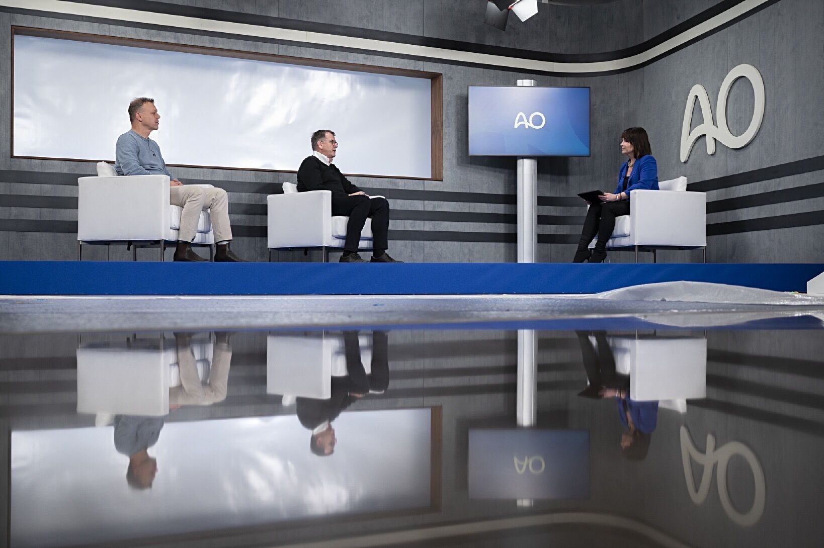 Three people sitting on stage chairs during a discussion in a modern studio, with a large screen displaying 'AO' behind them and a reflection of the scene on a highly polished floor.