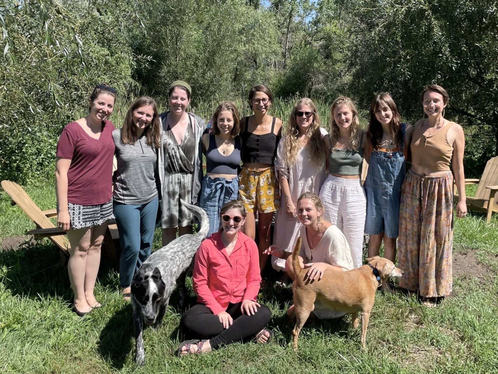 A group of 13 women and 2 dogs posing outdoors in a grassy area with trees in the background.