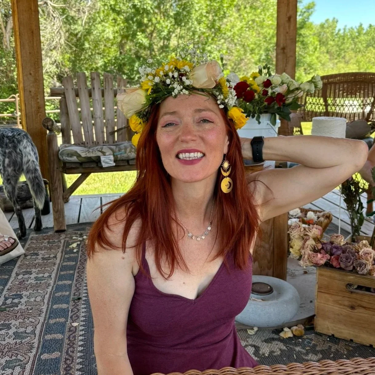 A woman with red hair wearing a floral crown, purple top, and yellow earrings smiling, in a rustic outdoor setting with flowers and greenery.