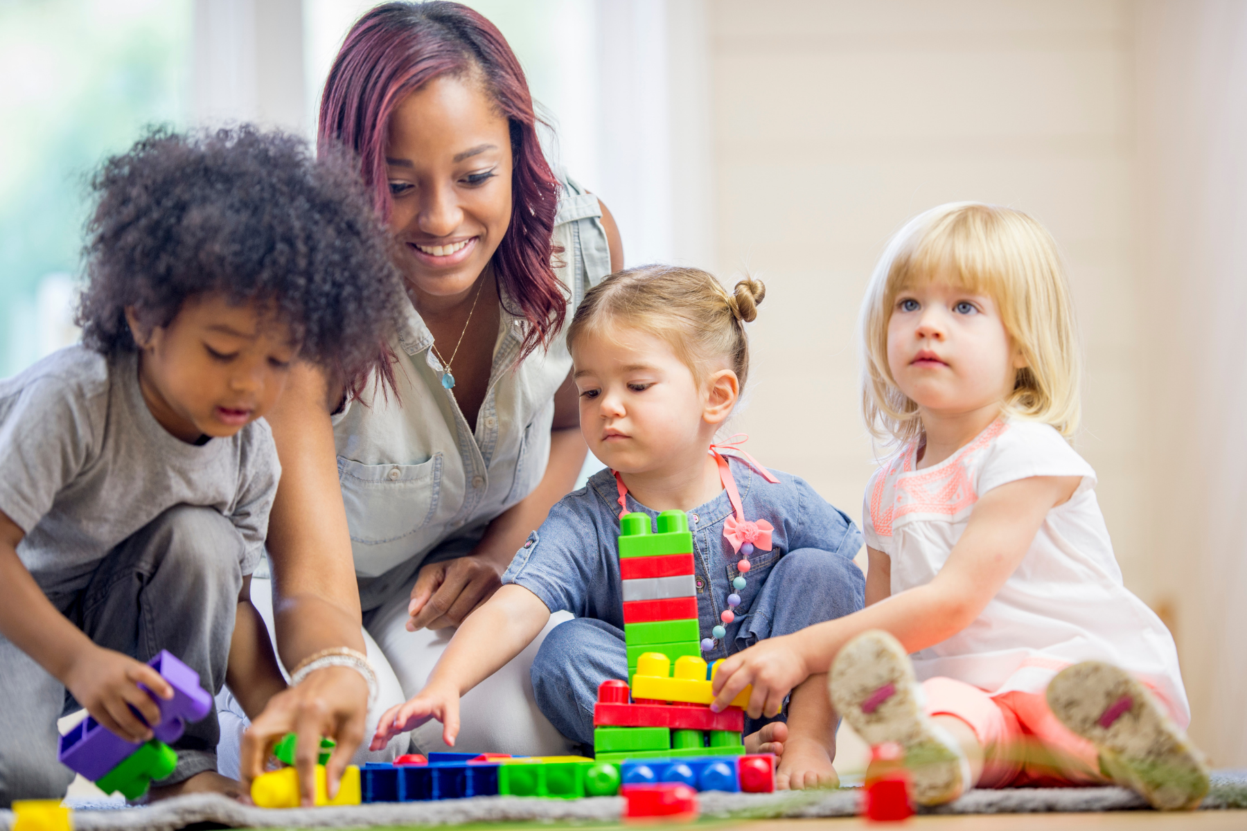 A young woman with red hair and a white shirt is playing with three young children and colorful building blocks on a rug indoors.