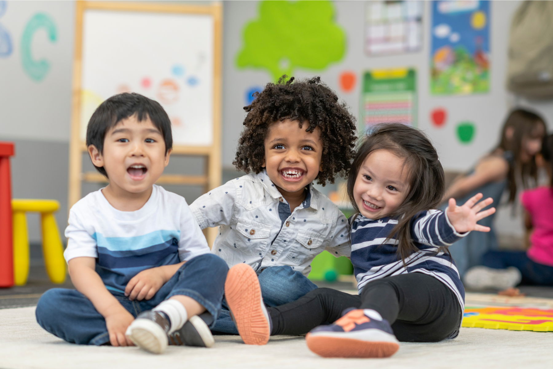 Three young children sitting on the floor in a classroom, smiling and playing together, with colorful educational posters and a whiteboard in the background.