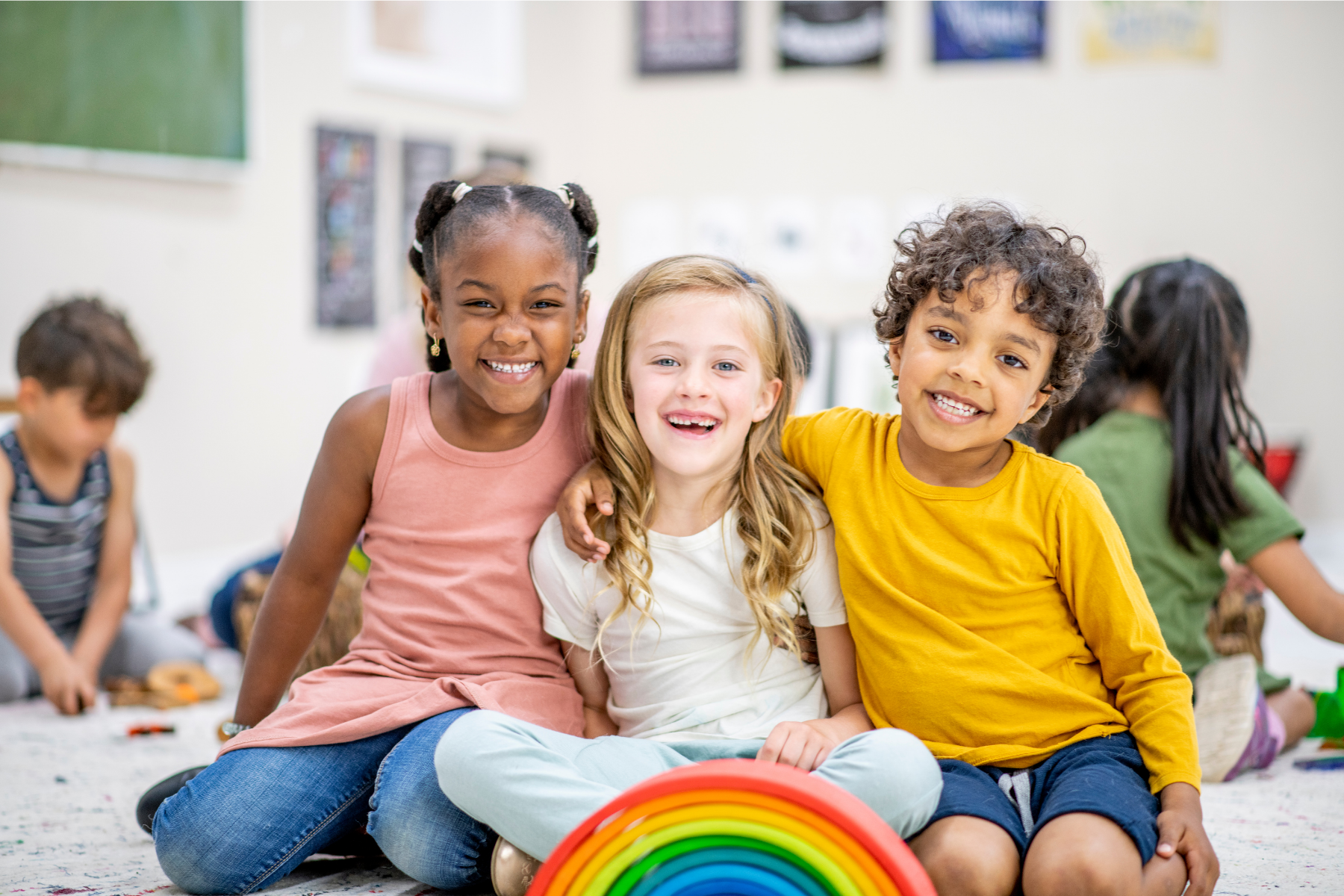 Three smiling children sitting on the floor in a classroom, with a rainbow toy in front, and other children in the background.