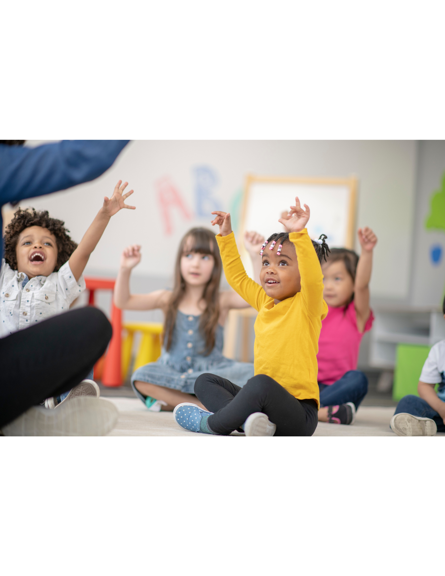 Young children sitting on the floor in a classroom, raising their hands and engaging with a teacher or instructor, with colorful decorations and letters on the wall.