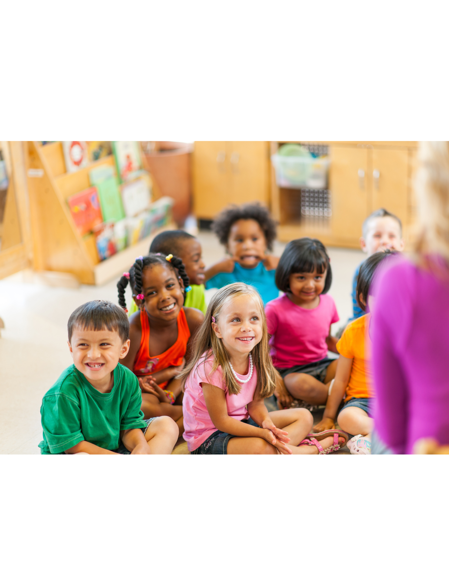 A group of smiling children sitting on the floor in a classroom, listening to a person in a purple top.