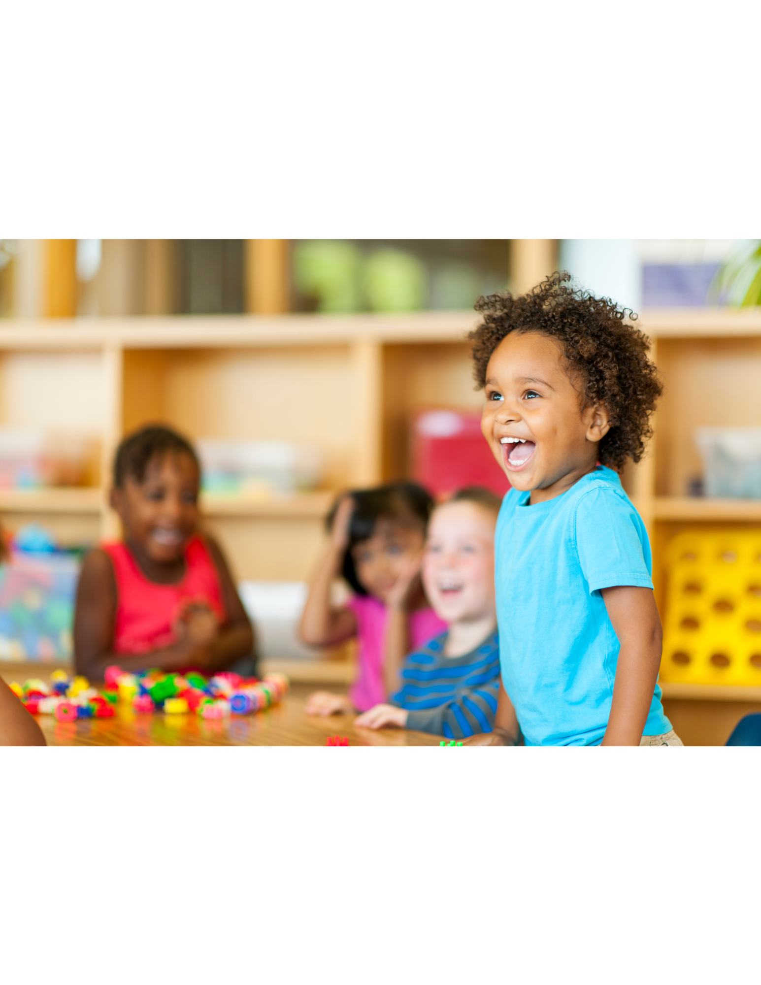 A group of young children sitting at a table, with one child in a blue shirt standing and smiling, while the others laugh and play with colorful toys in a classroom setting.
