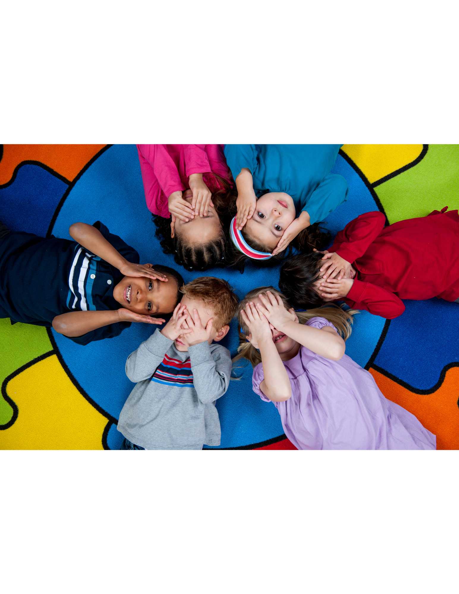 Six children lying in a circle on a colorful rug, covering their eyes with their hands and looking up.