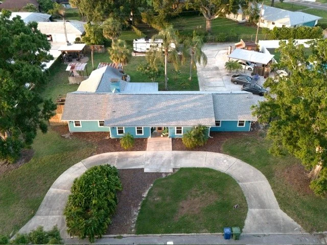 Aerial view of a single-story house with a curved concrete driveway, front lawn, surrounding trees, and neighboring houses.