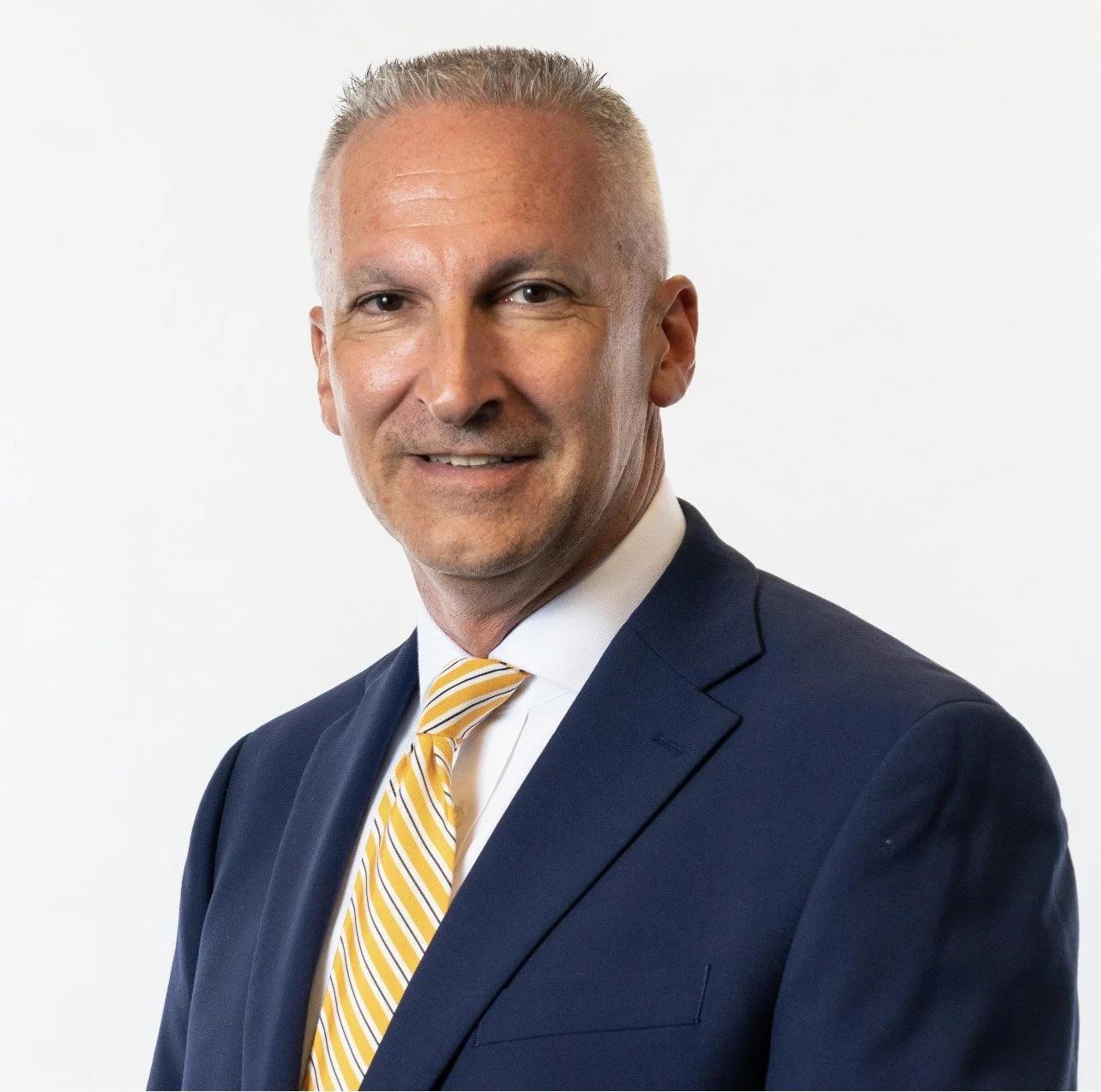Todd Metro headshot, wearing a dark blue suit, white shirt, and yellow striped tie, smiling slightly against a plain white background.