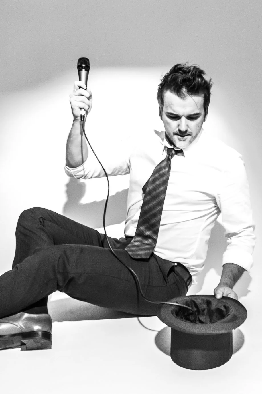 Ein Mann in Business-Kleidung sitzt auf dem Boden, hält ein Mikrofon in der Hand, ein Zylinder mit Zylinderhut vor ihm, und schaut nach unten.