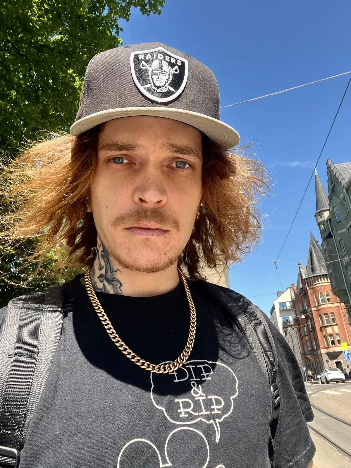 Nuori mies ottaa selfie, hänellä on raidallinen t-paita, ketju ja lämminhattu, jossa on Raiders-logo. Taustalla on kaupungin katu ja rakennuksia, sekä vihreä puu ja sininen taivas.