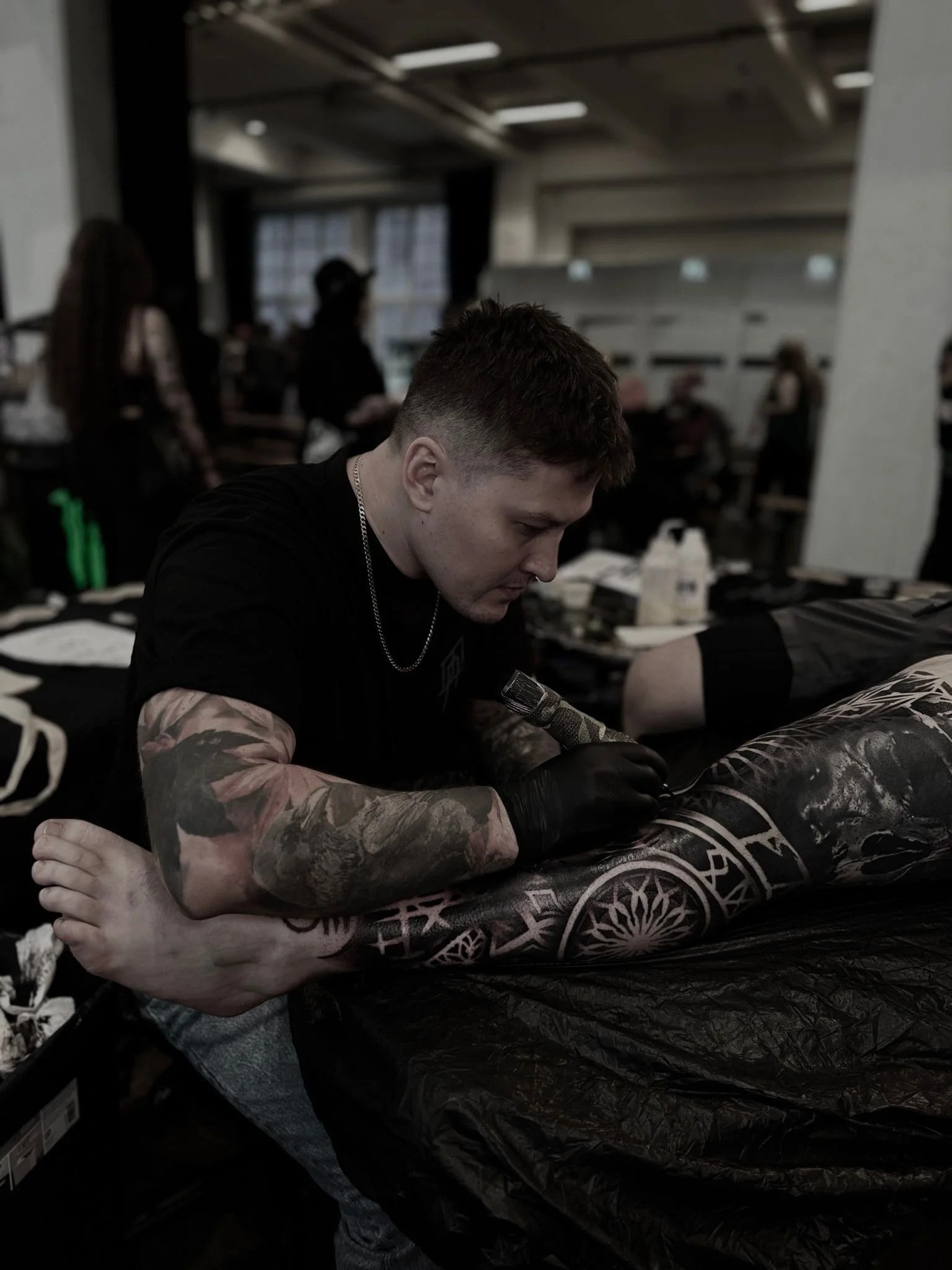 Kuvassa henkilö, jolla on tattooidut käsivarren ja jalkojen osat, makaa selällään pöydällä ja näyttää saavan tatuointia, ympärillä muita ihmisiä ja tatuointitarvikkeita.