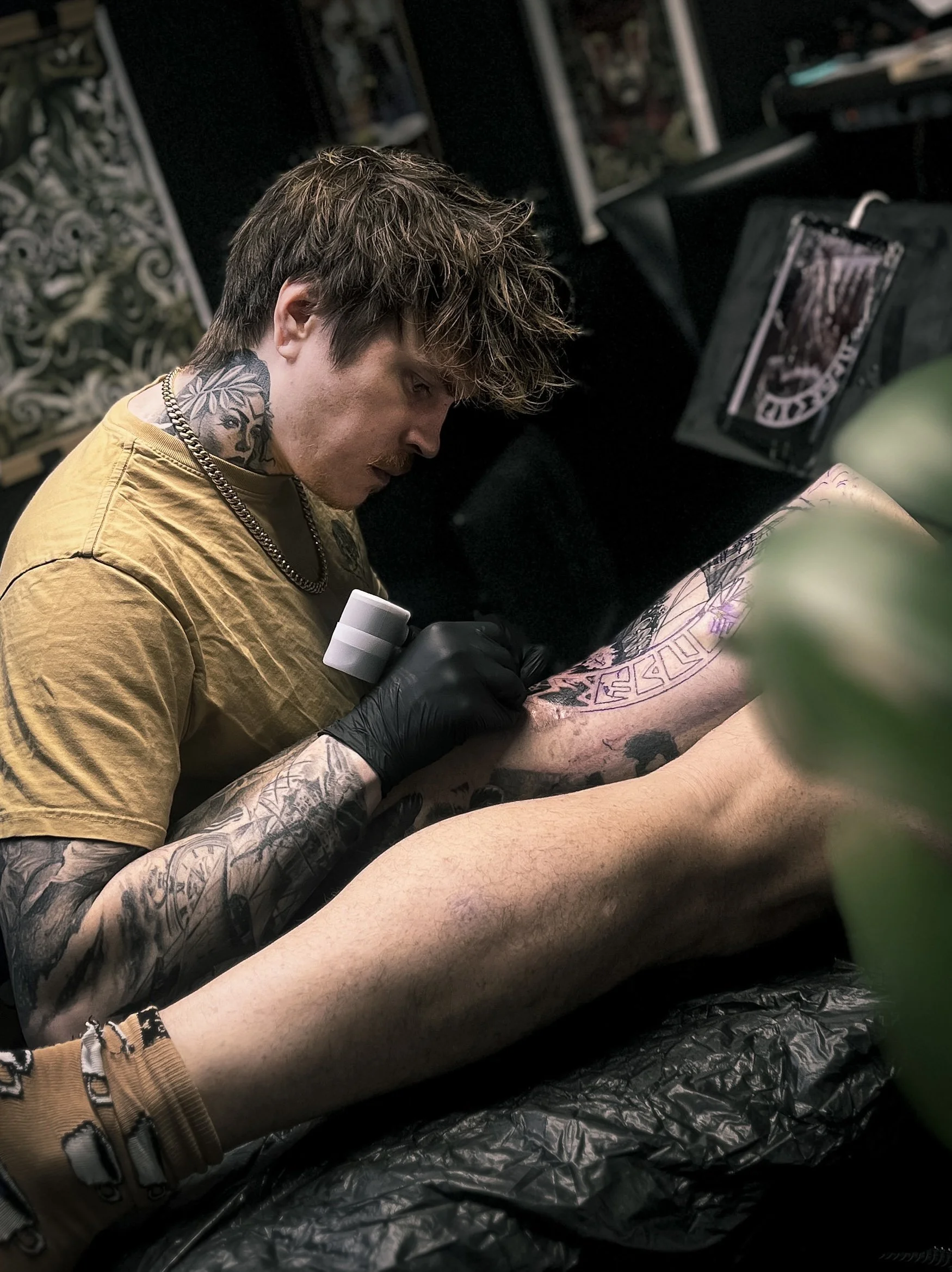Nuori mies, jolla on monia tatuointeja kaulassa ja käsivarsiensa yläosassa, työskentelee kehon tatuoinnin parissa tatuointistudiossa. Hän käyttää mustia käsineitä ja on keskittynyt työhönsä, joka sisältää kuvion tatuoimista toisen henkilön ihoon.