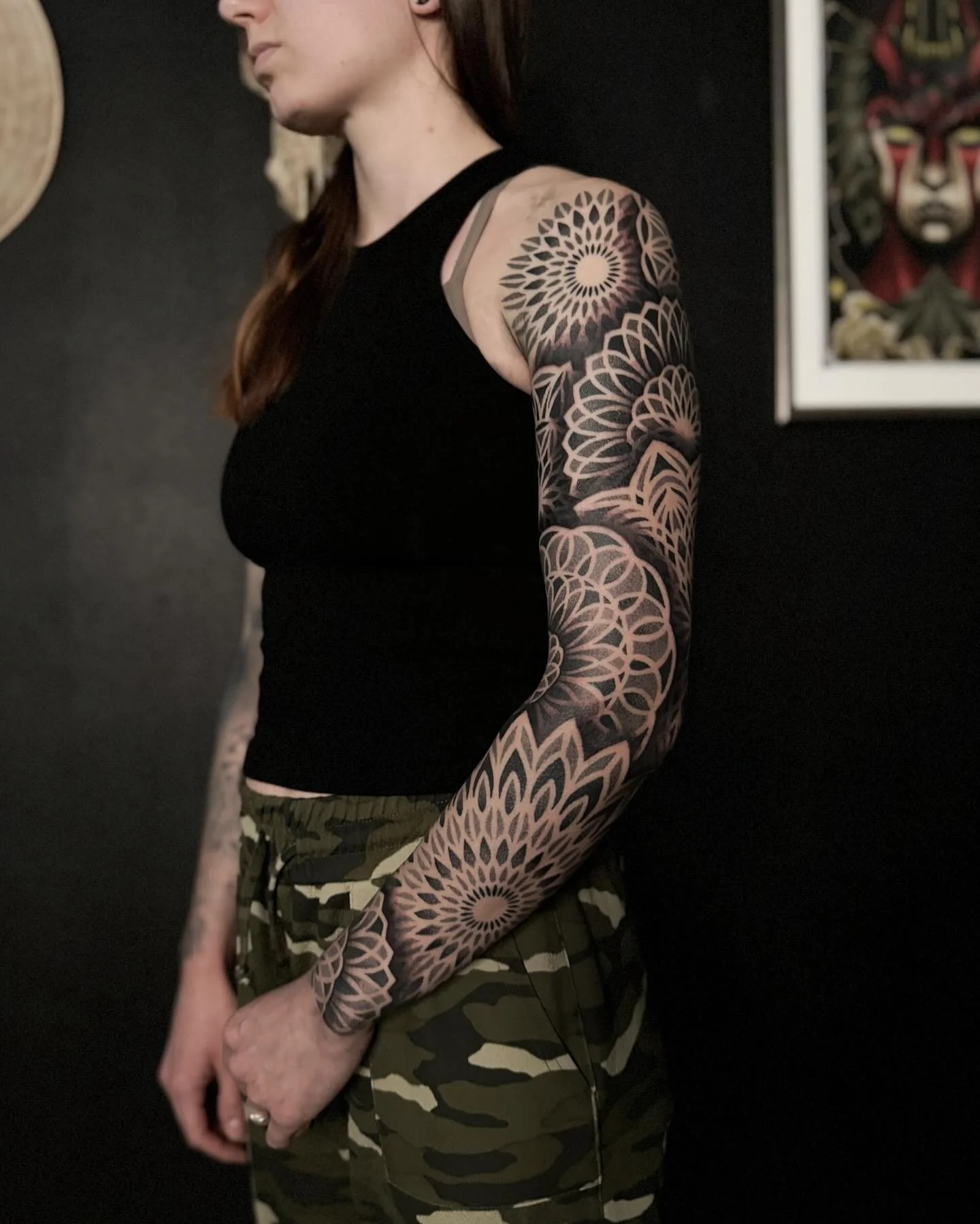 Kuvassa henkilö, jolla on pitkät hiukset ja selkä näkyvissä, hänellä on monimutkainen mustavalkoinen tatuointi oikeassa käsivarressaan, hän pukeutuu mustaan toppiin ja camouflage-housuihin.