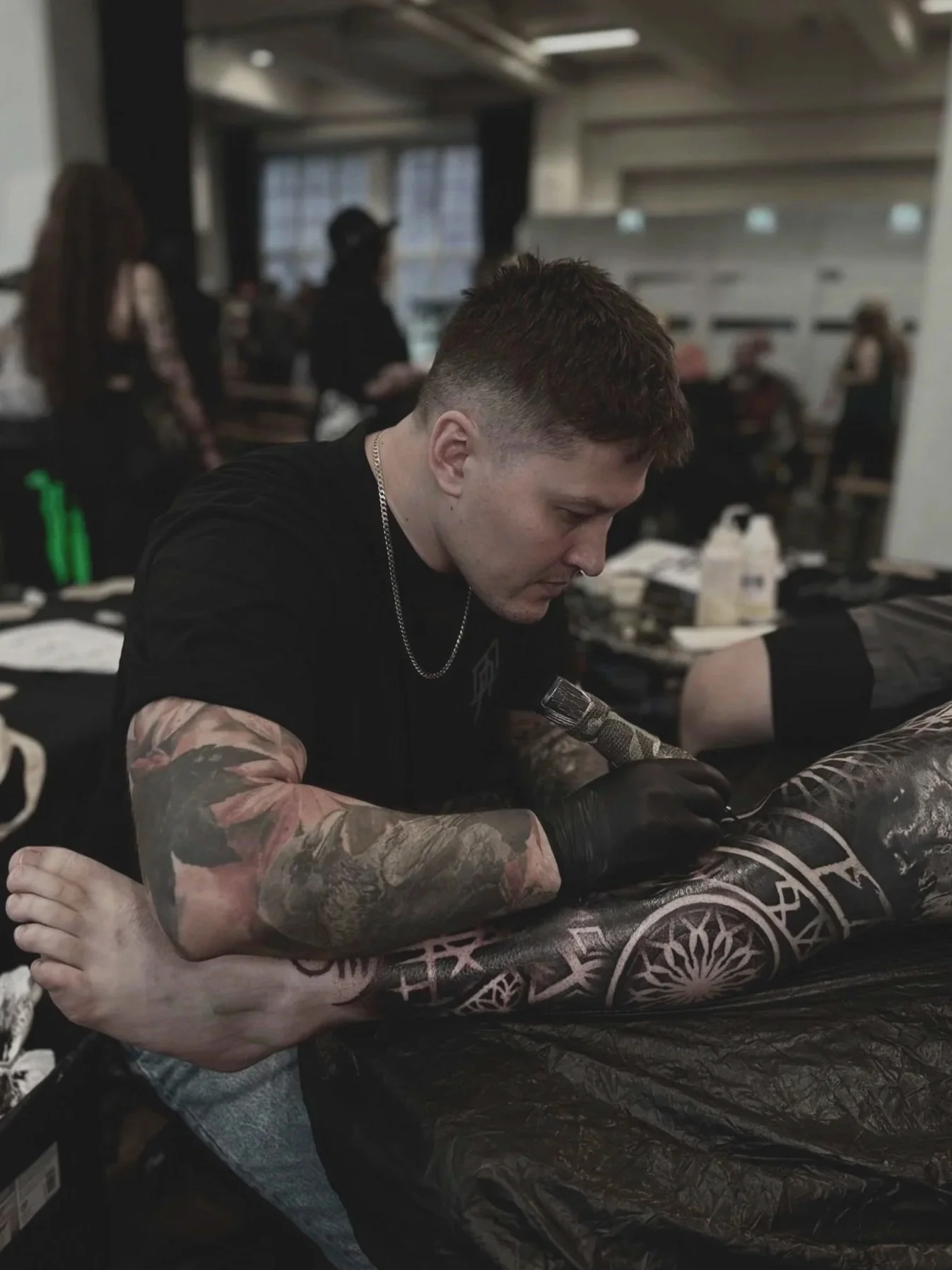 Nuori mies tatuoija tekee tatuointia asiakkaalle studiossa. Hänellä on tatuointimallit käsivarsissaan, ja sijaitsee jaetussa tilassa, jossa muita ihmisiä työskentelee taustalla.