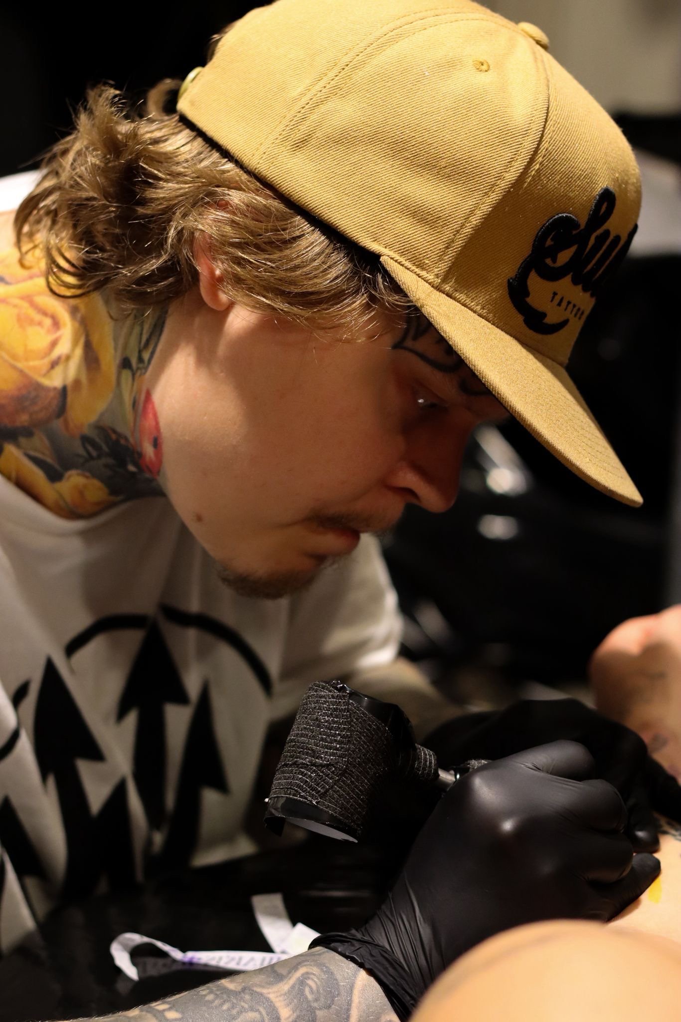 Henkilö pukeutuneena keltaiseen lippalakkiin, joka lukee 'Shawn' ja tattoo naamassa, istuu mustassa tuolissa ja katsoo alaspäin, mahdollisesti puhelinta tai muuta laitetta.