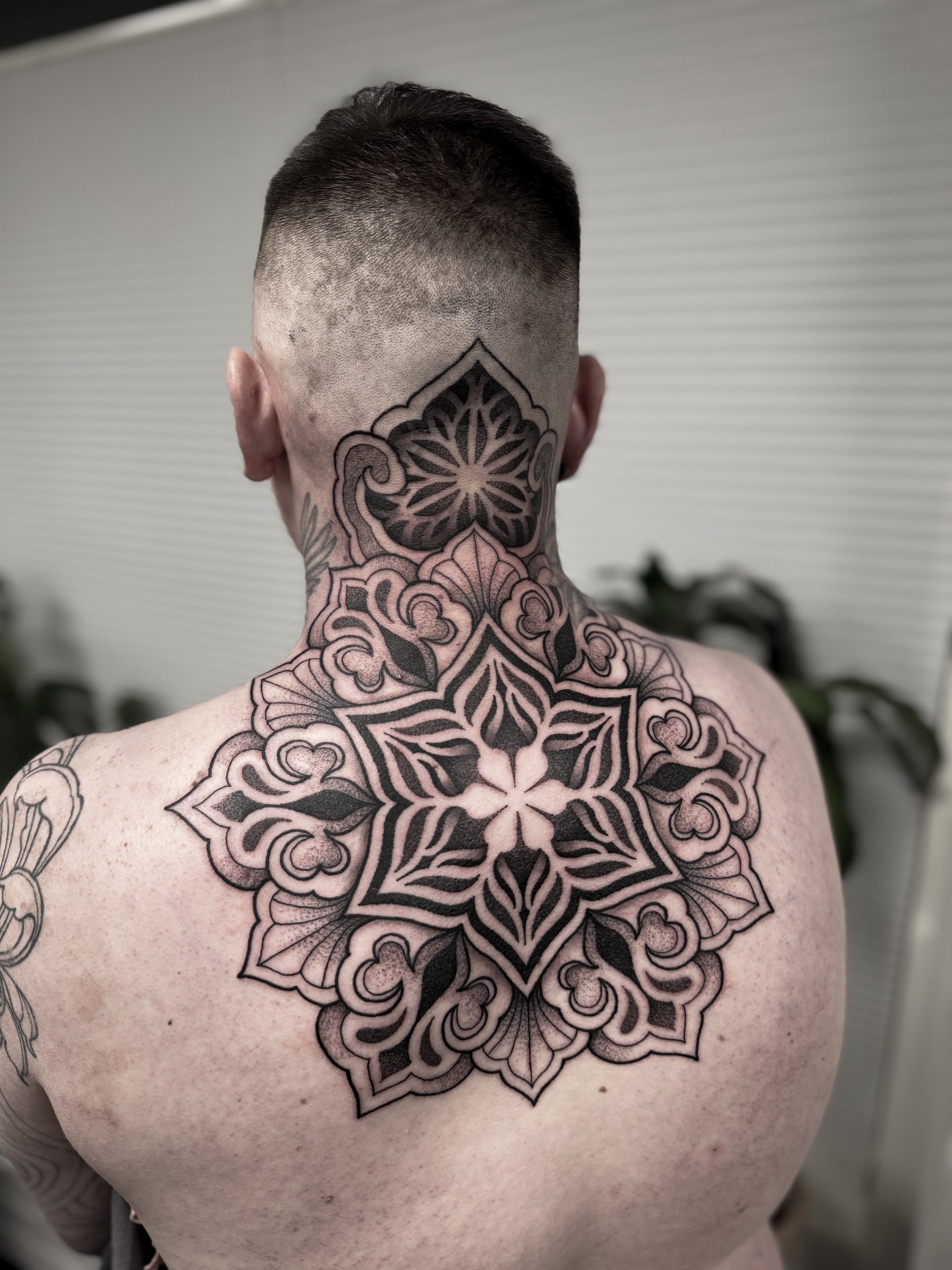 Henkilön selkä, jossa suuri mustavalkoinen tatuointi, joka sisältää monimutkaisen kukkamaisen kuvion. Taustalla vaaleanvärinen muovipaneli.