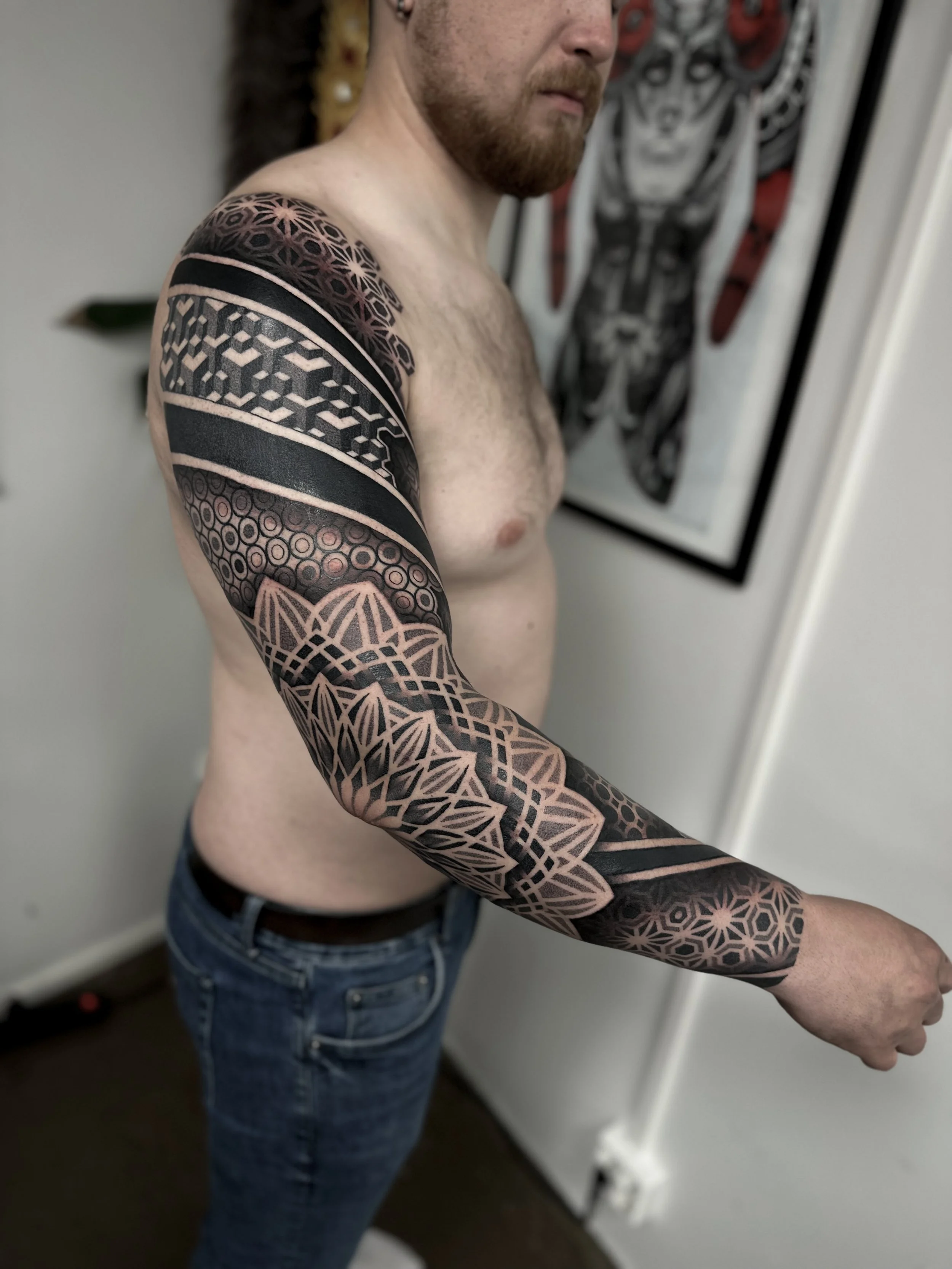 Nainen näyttää vasen olkapää ja oikea käsivarsi, jotka on tatuoitu geometrisilla ja graafisilla kuvioilla. Taustalla on seinällä taideteos ja ovi.