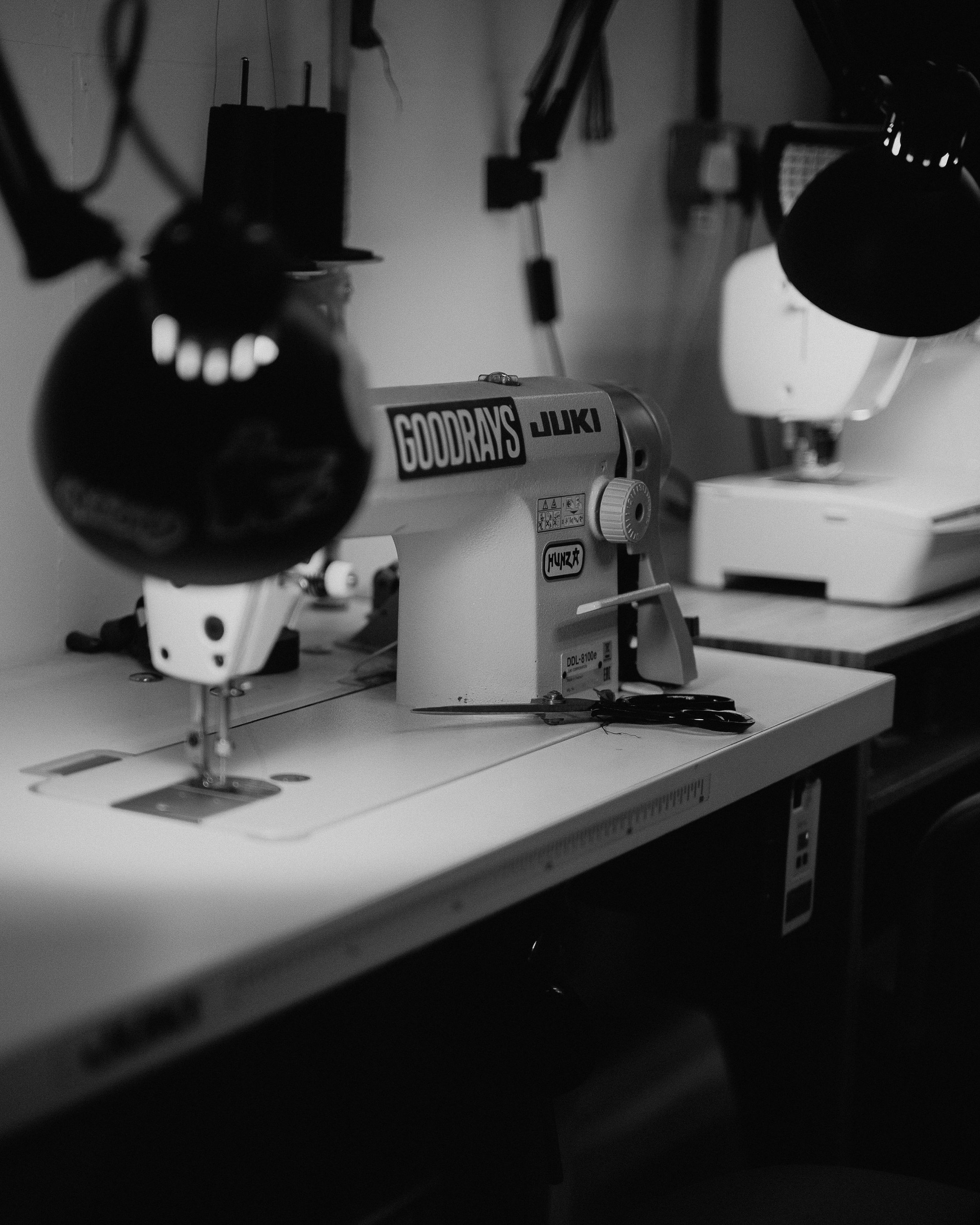 A black-and-white photo of a sewing machine on a worktable, with scissors placed on the table and sewing machine accessories in the background.